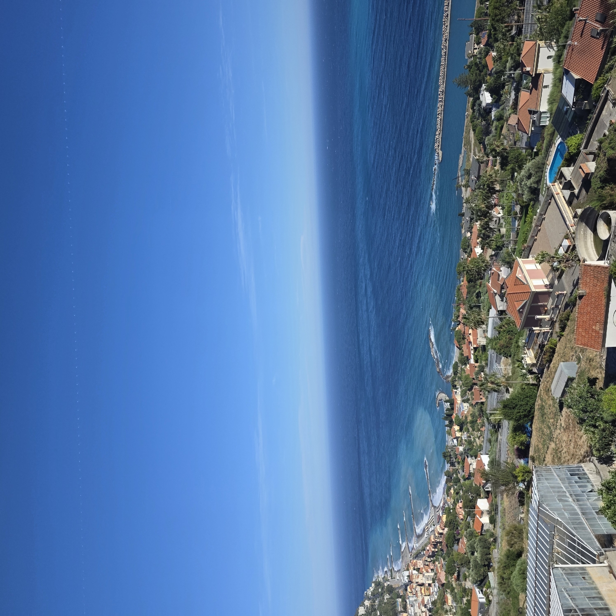The image shows a panoramic view of the coastal town of Ospedaletti, Italy, with the Mediterranean Sea in the background. The sky is a clear, vibrant blue, with a faint, dotted line of what appears to be contrails from an aircraft stretching horizontally across the upper portion of the frame.
The foreground features a greenhouse structure with a glass roof and metal frame, situated on a dry, grassy hillside. Beyond the greenhouse, the landscape descends towards the town, revealing a mosaic of terracotta-tiled roofs on houses and buildings scattered amongst lush green trees and vegetation. The town itself hugs the coastline, with a crescent-shaped beach and a series of wave-breaking structures extending into the turquoise and deep blue waters of the sea. A long, man-made breakwater, constructed of large rocks, extends out into the sea.
Further inland, a swimming pool with bright blue water is visible, surrounded by paved patios and a low wall. Various residential buildings with the characteristic red-tiled roofs of the region are nestled into the hillside, some appearing quite substantial with balconies and terraces. A road runs parallel to the coastline, with some vehicles visible. The overall impression is a sunny day in a picturesque Italian coastal town, with evidence of both residential life and agricultural activity (the greenhouse). There are no people visible in the image.