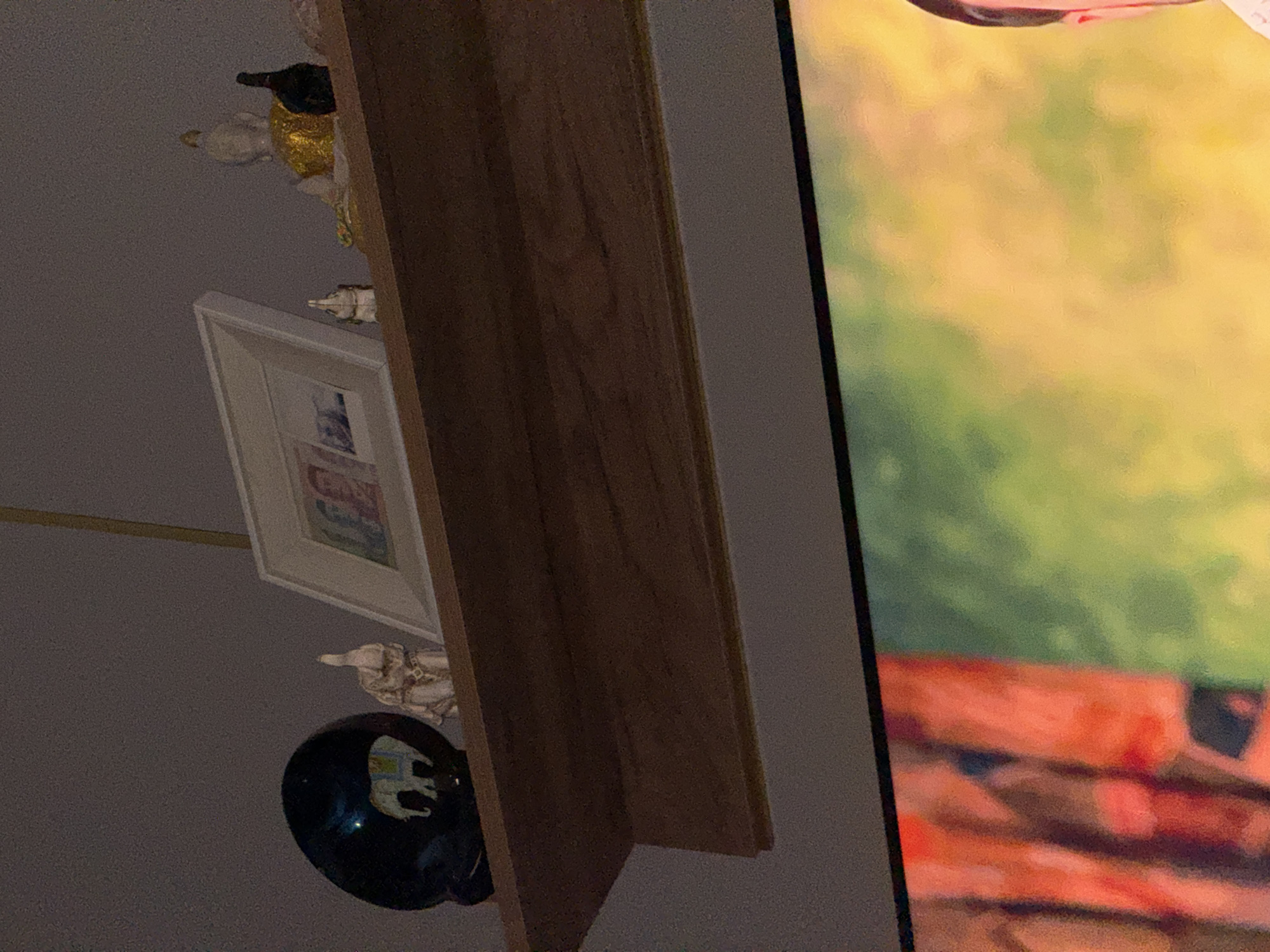A shelf above a television displays several decorative items. On the left is a dark, rounded object with a painted elephant design. Next to it is a small, light-colored elephant figurine. To the right of the elephant figurine is a framed picture. Further to the right is a seated Buddha statue with a gold robe and a dark object next to it. The shelf is made of wood. The image is from Mikhaylovsk, Russia.