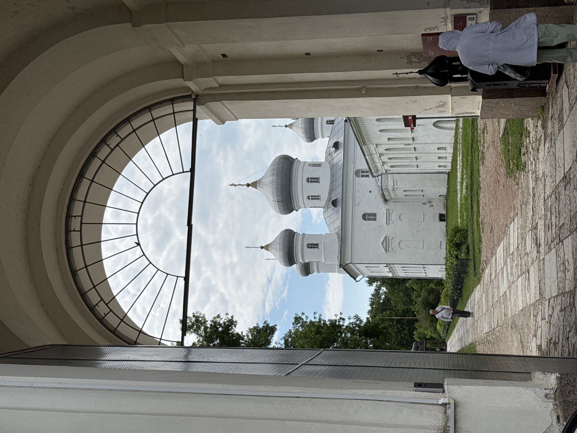The image shows an exterior view of a church with multiple domes, possibly part of the Novgorod Kremlin in Veliky Novgorod, Russia. The church is white with grey domes and crosses. In the foreground, an arched gateway frames the view of the church. A paved walkway leads towards the church, with a person walking away from the camera on the left side of the walkway, and another person wearing a hooded jacket standing near a black, church-like structure on the right side of the walkway. Trees and green grass surround the church and walkway. The sky is cloudy with some patches of blue.