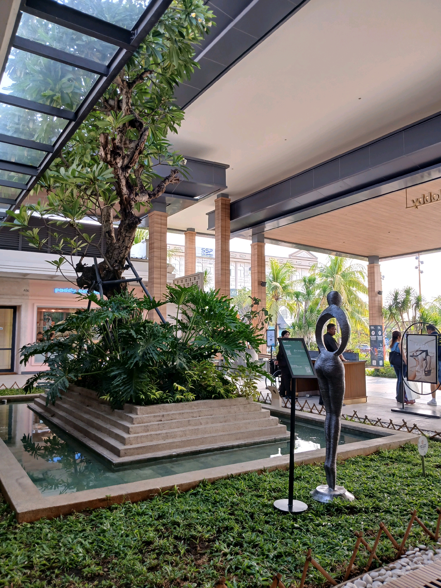An outdoor courtyard of a commercial complex in North Jakarta, Indonesia features a central water feature with tiered concrete steps leading into a shallow pool. Lush green plants, including large-leafed philodendrons and a small tree with support structures, surround the water feature. A black abstract sculpture of a human figure stands on a metal base in the foreground, positioned to the right of the water feature. In the background, several individuals are visible. Two individuals, one wearing a black hijab, appear to be near an information stand. Further back, a man is visible behind a counter, and two women are standing. The architecture of the complex includes a covered walkway with a sloped, transparent roof and wooden-paneled ceiling. The environment is well-lit, suggesting daytime.