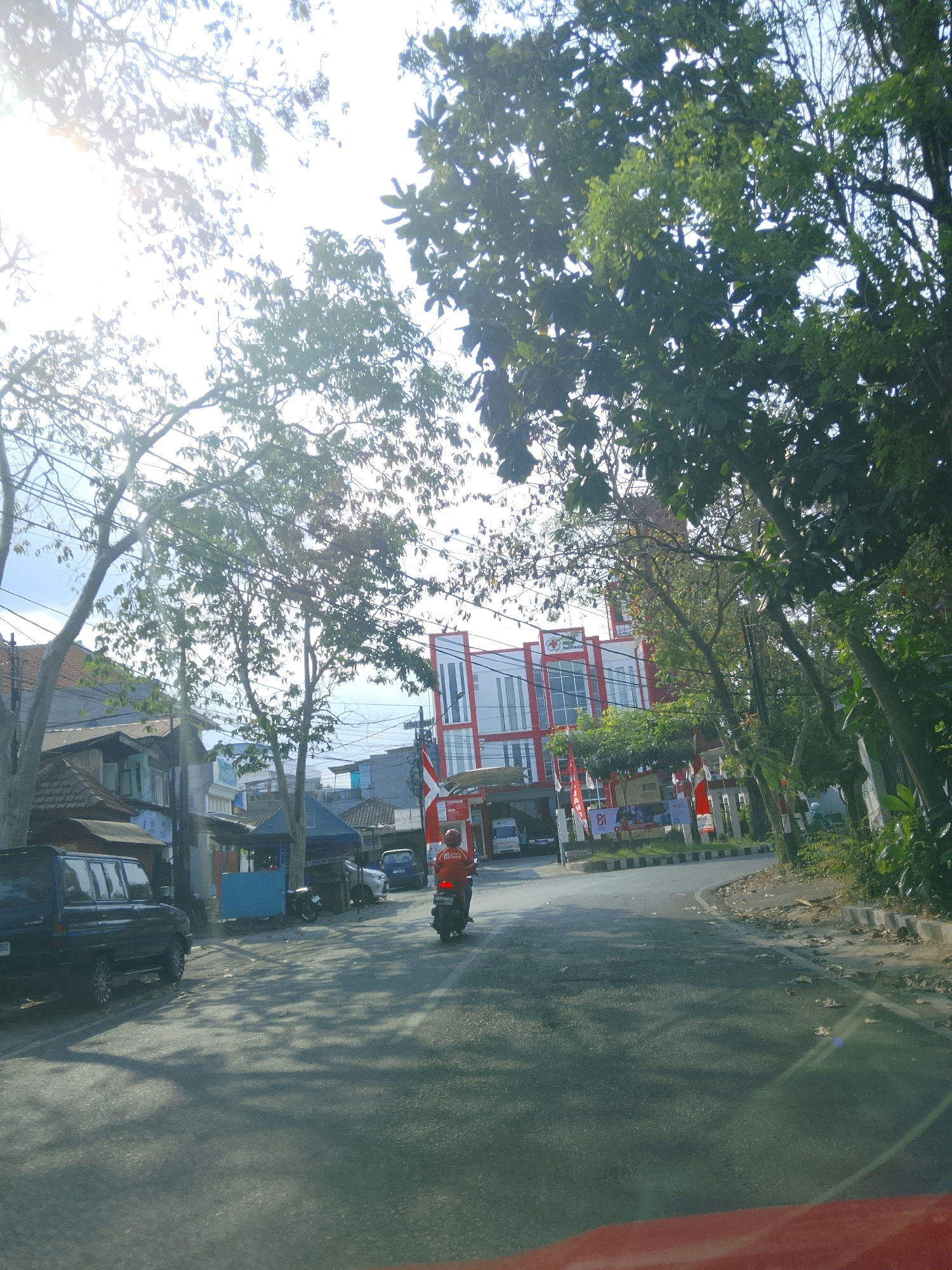 In Malang, Indonesia, a road curves past a modern, red and white building with large windows and a prominent logo that reads "Mitra Husada Indonesia". Several flags, some bearing the Indonesian flag, are displayed along the street. Trees with dense foliage cast shadows on the asphalt road. A person wearing an orange shirt is riding a motorcycle away from the camera. To the left, a dark-colored SUV is parked, and a smaller blue stall is visible further down the street. The sky is bright, with sunlight filtering through the trees.
