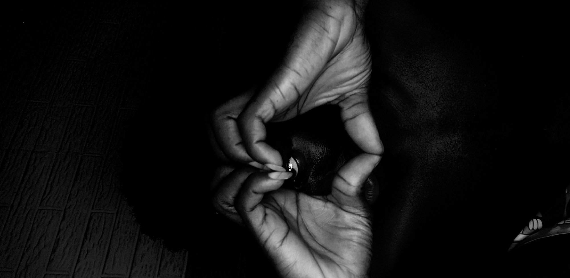 A black and white, close-up photograph shows a hand holding what appears to be a small black object in front of a reflective surface. The hand and its reflection are the primary subjects. The background is dark, but a surface with a pattern of rectangular shapes, possibly tiles, is visible to the left of the hand. The location, Narayi, Nigeria is not verifiable by the image alone.