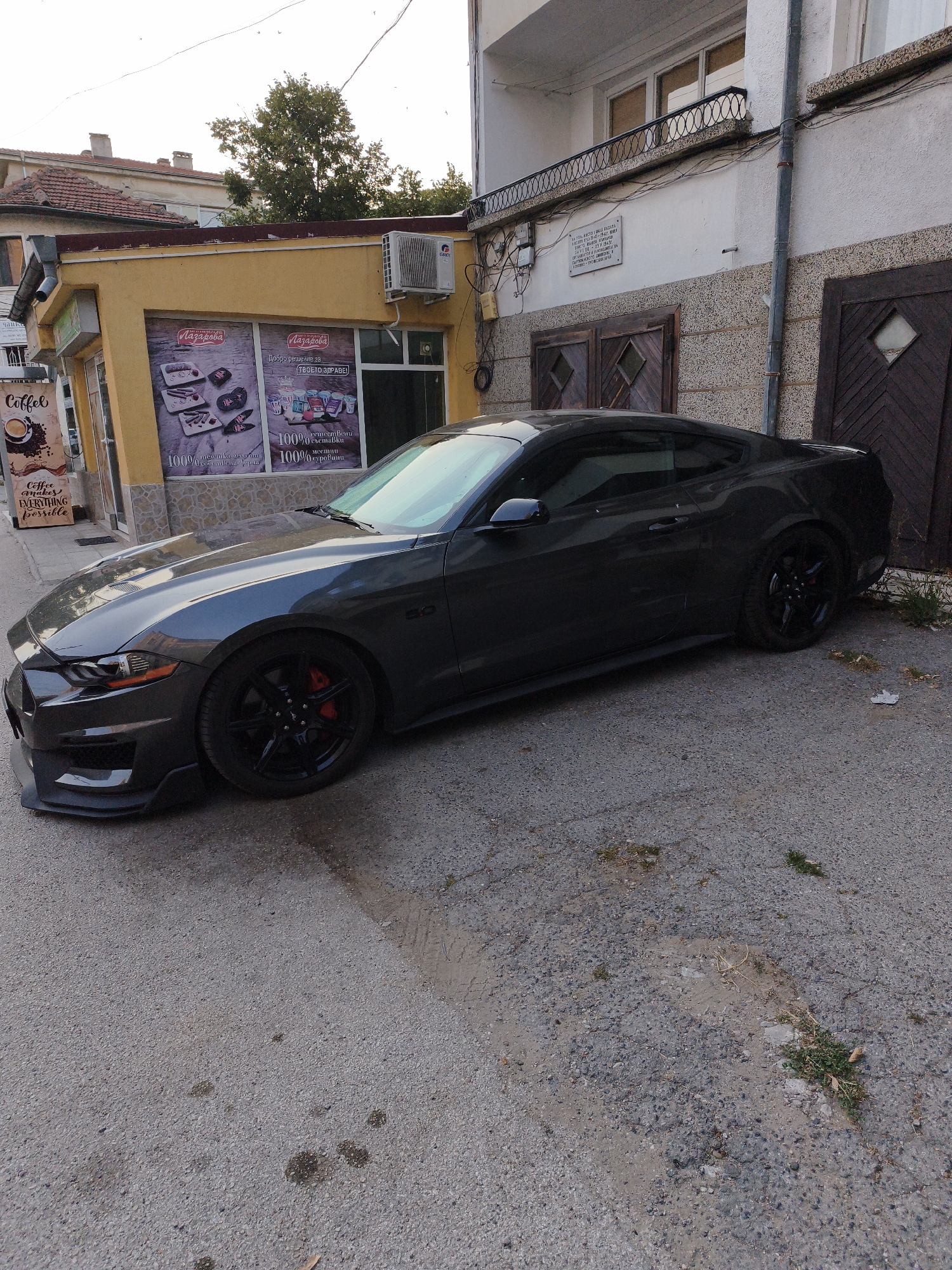 In Lovech, Bulgaria, a dark grey Ford Mustang is parked on a cracked asphalt surface in front of a yellow building with large display windows. The building appears to be a shop, with signage visible in Bulgarian. To the left of the Mustang, a vertical sign with "Coffee" written on it in English is mounted on the wall. The building to the right of the shop is white and grey, with wooden doors and a metal railing on an upper level. The overall scene depicts an urban street with a focus on the parked car and the surrounding architecture.
