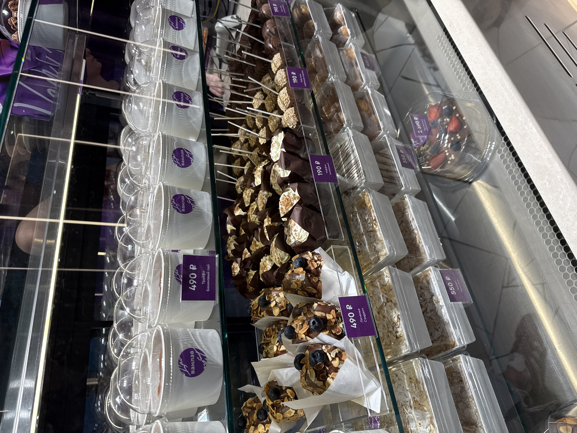 This is a close-up, commercial photograph taken indoors, likely in a dessert shop or bakery in Krasnoyarsk, Russia, as indicated by the Cyrillic script on the price tags. The focus is on a refrigerated display case showcasing various desserts, predominantly arranged in neat rows and columns.
On the left, there are several clear plastic cups labeled with a purple tag that says "Lau ванилав." These cups appear to contain some kind of trifle, described as "Банановый пай" (Banana pie) on the attached "490 P" price tag. Adjacent to these are chocolate-covered treats topped with almonds and blueberries, packaged in white paper wrappers with a matching "490 P" price tag that states “Печенье Сэндвич.”
To the right, there are chocolate-covered cake pops with toppings, marked with "190 P" price tags, one labeled as "Маффин Тыквенный" (Pumpkin muffin). The display case continues with rows of rectangular clear plastic containers filled with what seems to be layered pastries or bars, with tags indicating prices such as "550 P" for what is described as "Трайфл Наполеон".
Further to the right, a clear plastic container holds a chocolate dessert with strawberries and blueberries, priced at "2590 P", and the display case concludes with more pastries priced at "650 P" and more cake pops priced at "190 P".
The environment is brightly lit, typical of commercial display cases designed to highlight the products. Reflections on the glass surface of the display case suggest the presence of other objects and light sources in the surrounding environment, but these details are difficult to discern due to the focus of the shot.