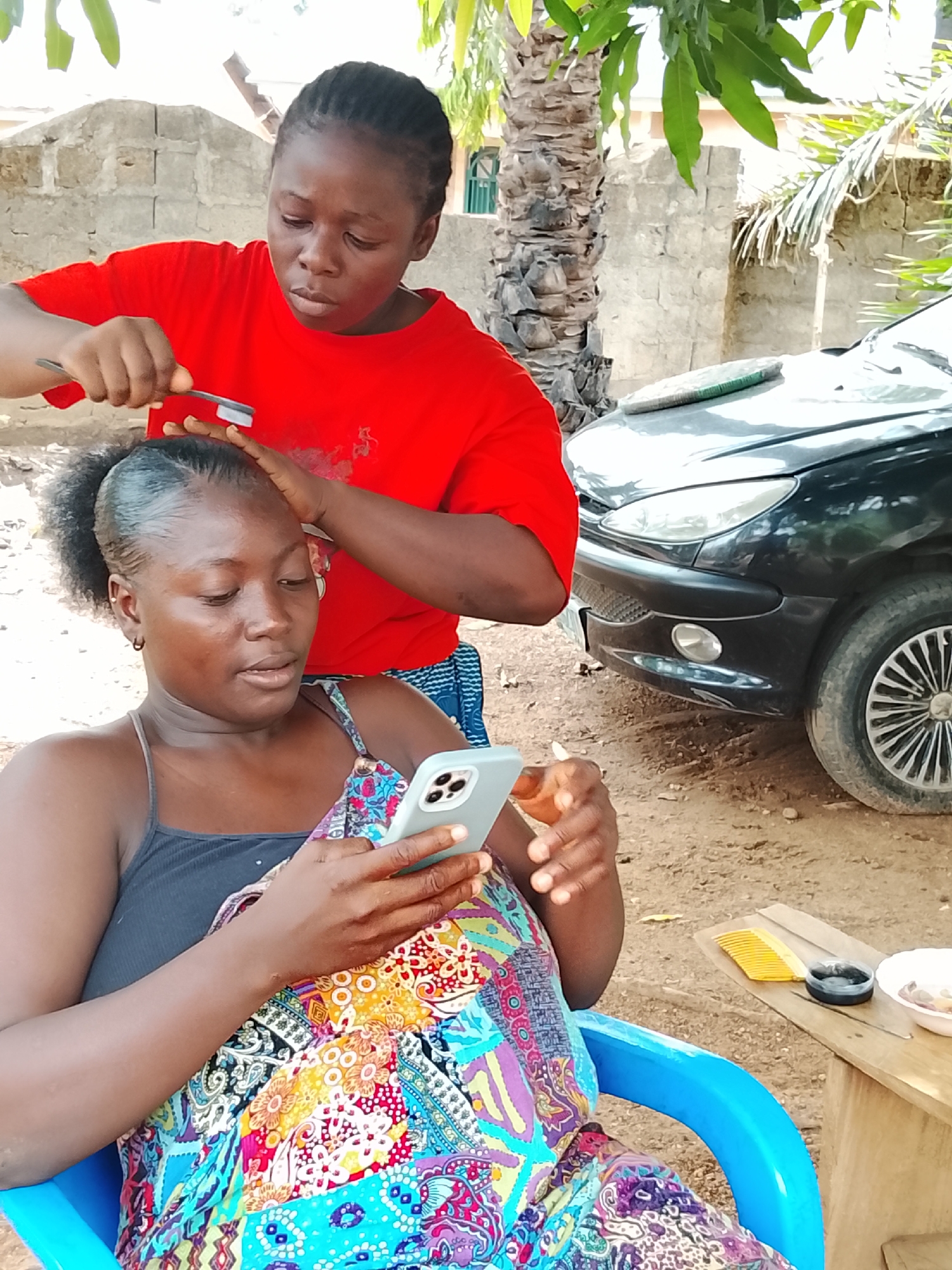 In Akwanga, Nigeria, a woman in a red shirt combs the hair of another woman seated in a blue plastic chair. The seated woman is looking at a mobile phone in her hands. A black car is parked in the background near a palm tree. A small wooden table is beside the seated woman, holding a comb, a small container, and a bowl. The setting appears to be outdoors, with a building visible in the background.