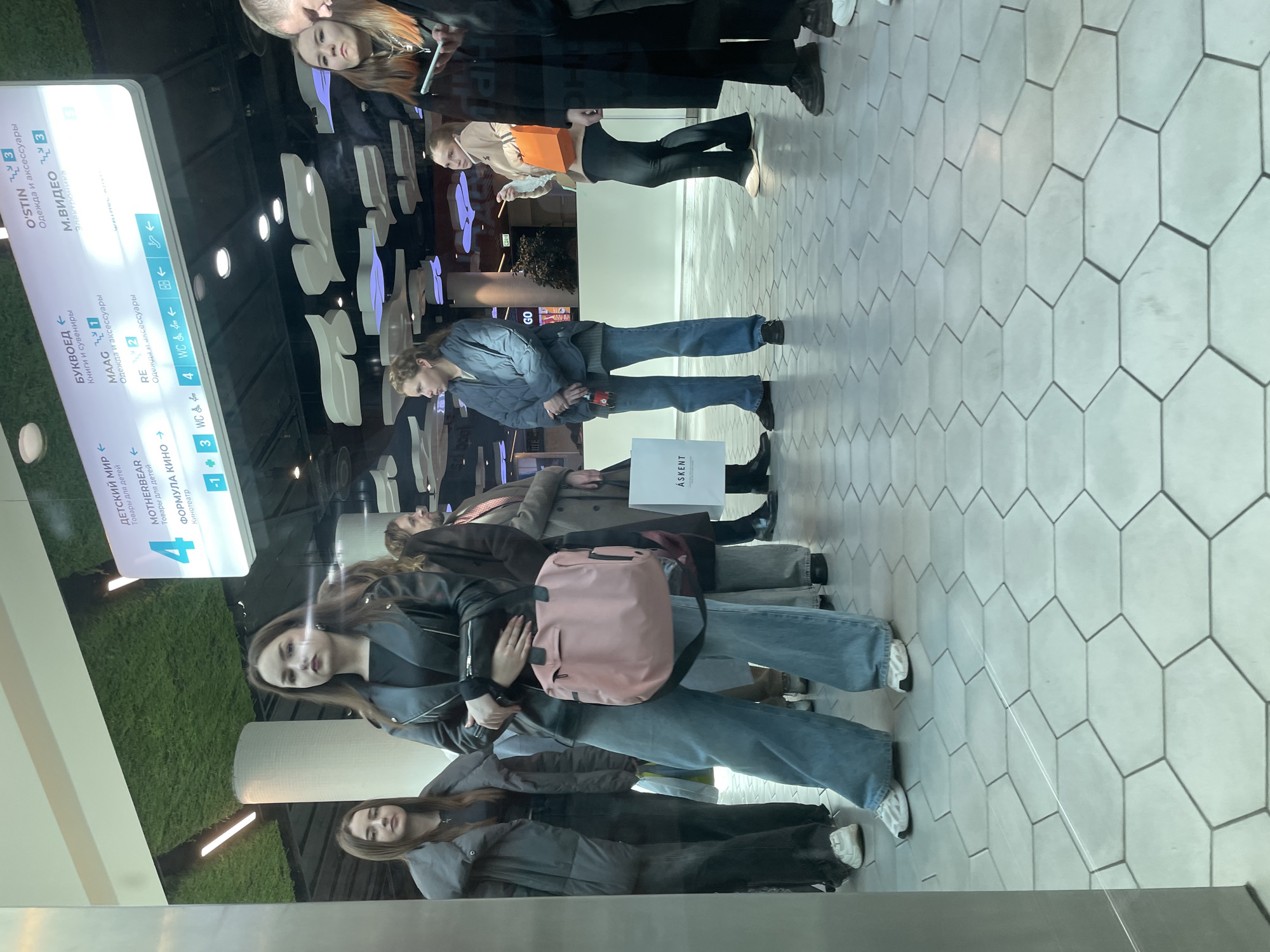 The image depicts a group of people reflected in a glass surface, likely a window or display case. The reflections suggest an interior setting, possibly a shopping mall or commercial area.
On the left side of the frame is a signpost with directions to various businesses. Some of the businesses listed are "O'STIN," "M.VIDEO," "BUKVOED," "MAAG," and "RE" with the text written in Russian. The sign uses a mix of text and directional arrows. The sign also indicates the presence of a "CHILDREN'S WORLD" and "FORMULA KINO" with floor numbers 4 and -1, respectively.
The people reflected in the glass vary in age and attire. Many are wearing jackets, jeans, and other casual clothing. One individual carries a pink bag. It appears the reflections are superimposed on a green background, potentially a display or artificial greenery within the mall. The floor is covered with a light gray hexagonal tiled floor.
Based on the businesses listed on the signpost and the reflection, it is likely that this scene is taking place in a shopping mall in Saint Petersburg, Russia.