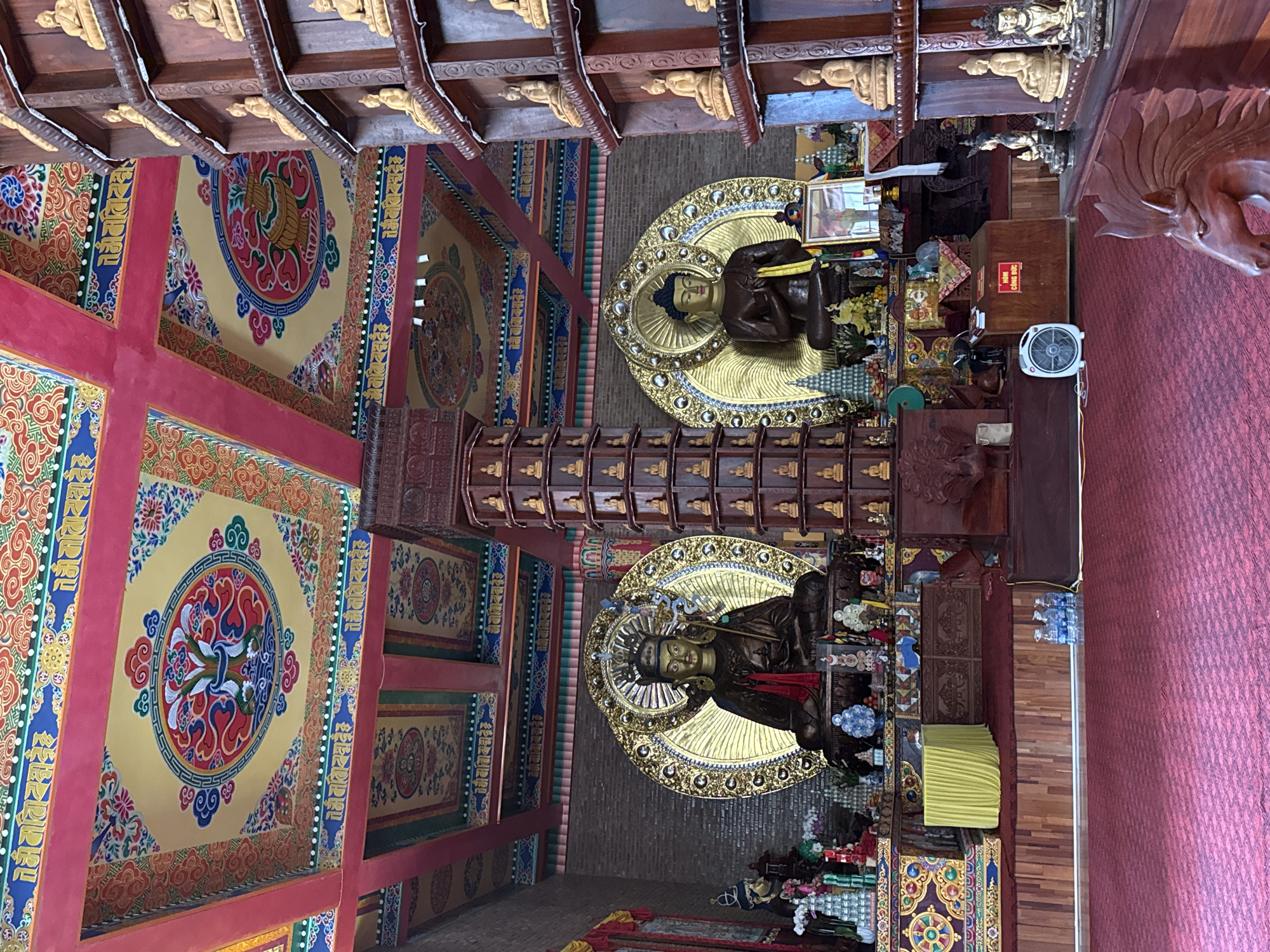 The image depicts the interior of a Buddhist temple in Việt Trì, Vietnam. The scene is dominated by two large, seated Buddha statues, one on the left and one on the right, each adorned with golden halos. In front of the statues, the floor is covered with a red carpet, and there is a wooden altar area with various offerings and decorations. To the right, a tall, wooden shelf structure is filled with numerous smaller Buddha figurines. The ceiling is elaborately decorated with colorful, intricate patterns and motifs. The overall atmosphere suggests a sacred and ornate religious space.