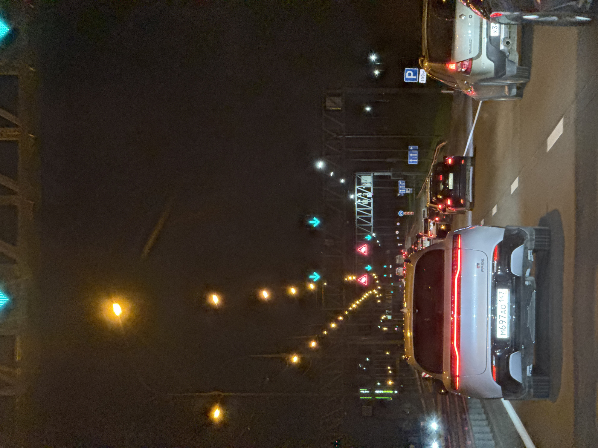 The image depicts a nighttime scene on a multi-lane highway in Vartemyagi, Russia. The road is illuminated by streetlights, traffic lights, and vehicle headlights. Several cars are visible, including a white SUV in the foreground with license plate M697AO 147. Other vehicles include a dark SUV, a silver hatchback, and a black sedan. Traffic lights are showing green arrows, indicating that lanes are open. Road signs, including a parking sign, are also present. The environment appears to be a typical urban or suburban highway setting at night. There are no people clearly visible in the image.