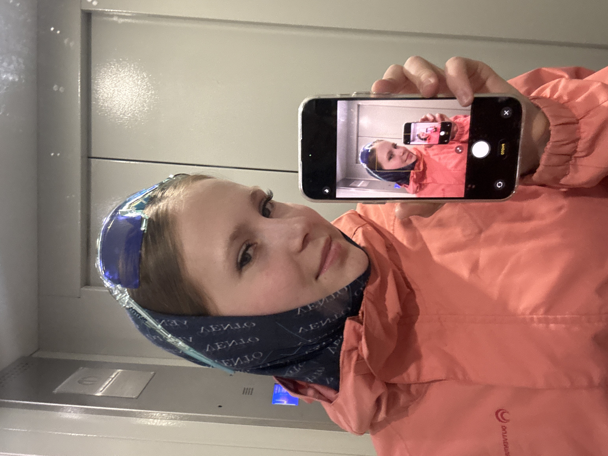 A woman in a coral-colored jacket and a dark blue headscarf with white text is taking a selfie in what appears to be an elevator. She's using her smartphone to capture the image, which is visible on the phone's screen, showing a smaller version of herself in the same attire and pose. The elevator's interior is a light gray color with a metal control panel visible on the left side of the frame. Given the prompt's request to identify the location and the association of the name "Chaikovsky" with a city in Russia, it's reasonable to assume this photo was taken in an elevator within Chaikovsky, Russia.
