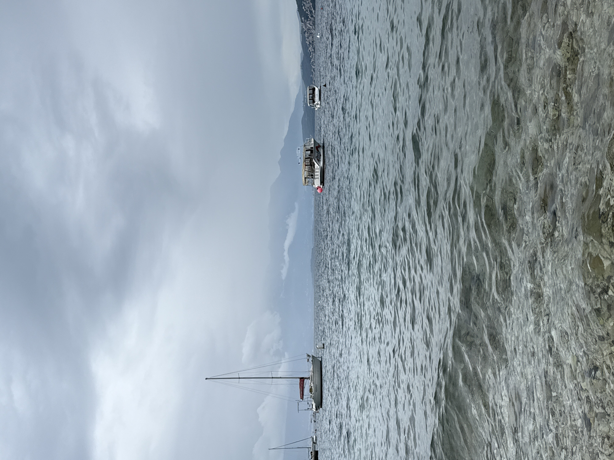 The image shows a body of water with several boats, including a sailboat with a red sail. In the distance, there is a mountain range. The sky is overcast with grey clouds. The water appears choppy and grey. Based on the presence of boats and a mountainous backdrop, the location is likely coastal. The location in the city of Đenovići, Montenegro cannot be verified with complete certainty from the image.