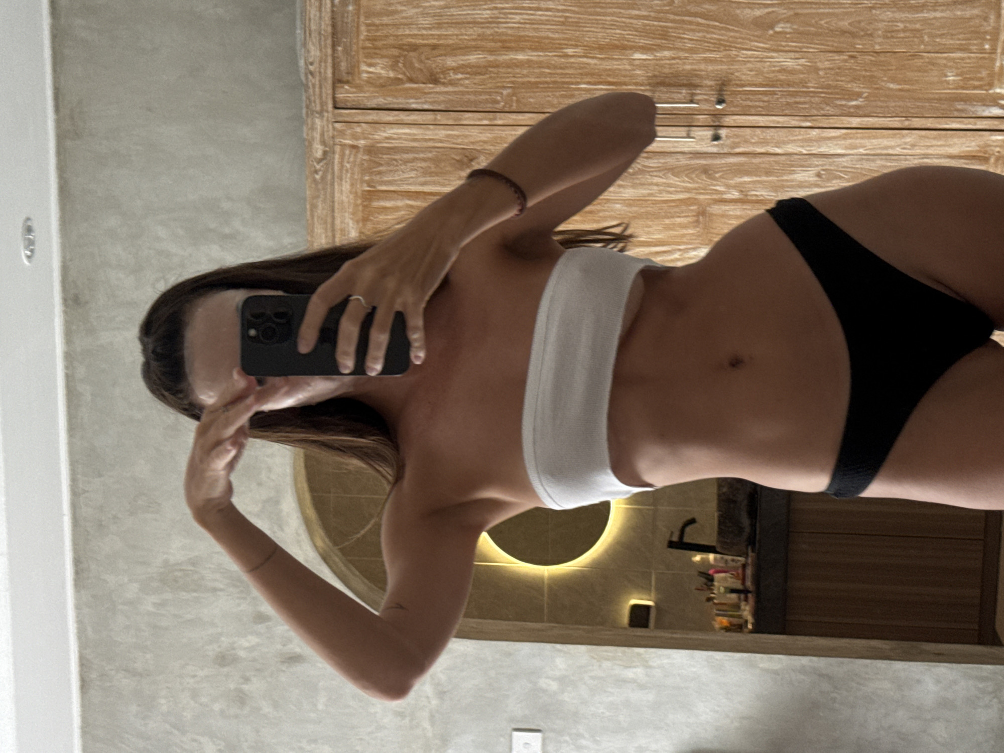 The image captures a mirror selfie of a person, likely female, in a bathroom setting in North Kuta, Indonesia. The person's face is mostly obscured by a black smartphone as they hold it up to take the photo. She is wearing a white bandeau top and black bikini bottoms or underwear, revealing her midsection. A ring is visible on her left hand, and a thin, dark bracelet is on her right wrist.
The background features a light-colored, possibly concrete or textured wall on the left, and a light-toned wooden cabinet or set of drawers with metal handles on the right. In the deeper background, a large circular mirror with warm backlighting illuminates the area, below which a dark faucet and various toiletries on a counter are reflected. The overall ambiance suggests a modern and well-lit bathroom. The image is oriented with the person lying on their side.