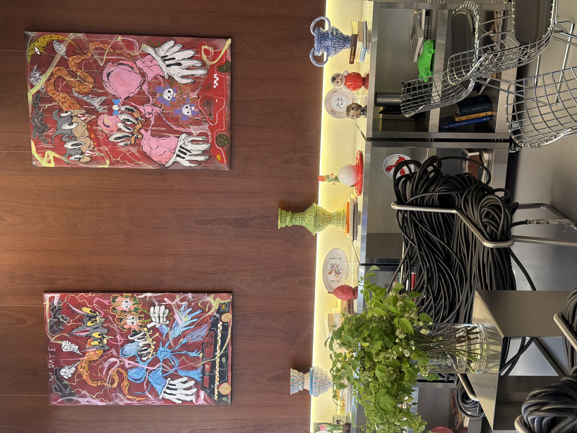 This image captures a close-up interior shot of a room, possibly a gallery or a well-decorated living space, featuring artwork and decorative objects. The scene is dominated by two vibrantly colored paintings hanging on a dark wooden paneled wall. Both paintings depict cartoonish figures, possibly inspired by classic animation characters, rendered in a somewhat chaotic and expressive style with bold lines and bright hues of red, pink, blue, and yellow. The painting on the left has the text "THE DATE" visible at the top.
Below the artwork, a sleek, modern metal shelving unit is visible, illuminated by a strip of warm yellow light. On the shelves are various decorative items, including ceramic vases of different shapes and colors. One vase on the left is a textured light blue and white, while a striking tall green vase with an ornate texture is placed to the right. Between these, there are smaller decorative pieces like a red pear-shaped object, a small plate with floral motifs, a small brown sculptural figure, and a red and white bobble doll. A blue ceramic vase with looped handles sits on the right side of the shelf.
In the foreground, a low, modern metal table holds a clear glass vase filled with lush green foliage and small white flowers. To the right of this table and partially in front of the shelving unit, a large quantity of coiled black cables is artfully arranged, creating a dramatic visual element. Further to the right, several metallic wireframe chairs with black seats are positioned. A bright green toy car is visible on the top shelf of the metal unit. The overall impression is one of a curated space that blends contemporary art, decorative accents, and industrial design elements. The image does not contain any identifiable landmarks or context that would allow for its specific location within Saint Petersburg, Russia, to be determined.