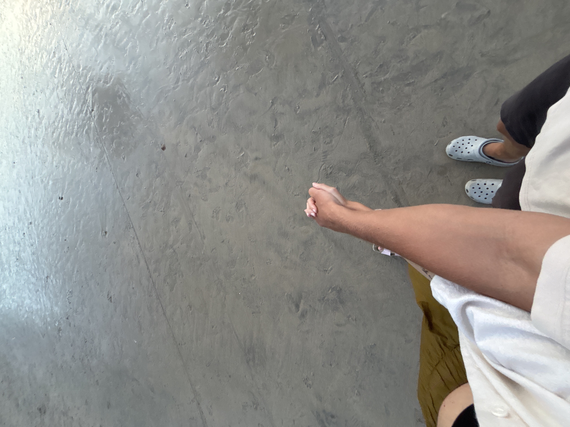 An overhead shot captures two individuals standing on a textured, light gray concrete or polished cement floor in Kuta Selatan, Indonesia. In the foreground, a person's arms and hands are visible, with the hands clasped together, suggesting a moment of waiting or introspection. This person is wearing a white shirt and appears to have a piece of olive or khaki-colored fabric (possibly pants or a skirt) below the shirt. Their skin tone is medium.
Further into the background, another person's feet are visible, wearing light blue Crocs shoes and dark-colored pants. The bright and even lighting, combined with the smooth floor, suggests an indoor setting or a covered outdoor area. No specific activity beyond standing and waiting is evident. No text is visible in the image.