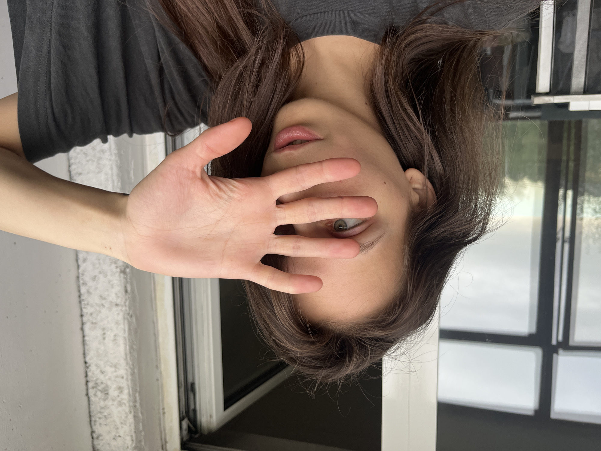 A woman with long, brown hair is holding her hand up, partially obscuring her face. Her right eye is visible between her fingers. She is wearing a gray t-shirt. The background shows a window and a blurry view of the outside. Given the available information, it is impossible to determine the specific location within Krasnoyarsk, Russia.