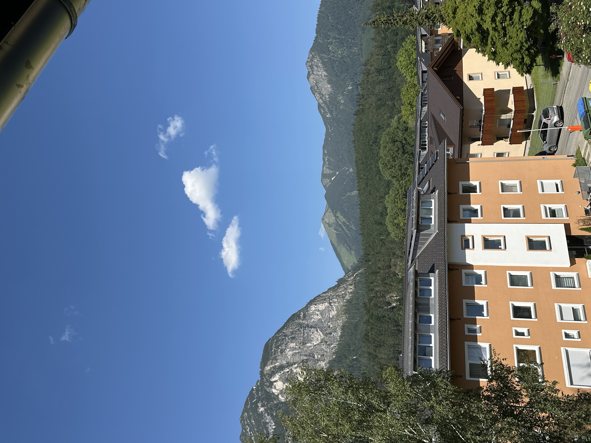 The image depicts a picturesque scene in Garmisch-Partenkirchen, Germany. The setting is a residential area with buildings nestled against a backdrop of lush green mountains. The time of day appears to be late morning or early afternoon, indicated by the bright sunlight casting shadows. The weather is clear and sunny with a vibrant blue sky dotted with a few fluffy white clouds.
In the foreground, a large section of a green downspout or pipe occupies the left edge of the frame, suggesting the viewpoint is from a balcony or window. The main subjects are several multi-story residential buildings, characterized by their warm, earthy tones like orange and cream. These buildings feature numerous windows, some with white frames and others with visible blinds or curtains. Balconies with wooden railings are also visible on some of the structures. The architecture suggests a modern European style.
Behind the buildings, the majestic mountains rise, covered in dense green forests with rocky outcrops and patches of snow visible higher up. The mountains are sharply defined against the clear sky, showcasing their impressive scale and natural beauty. Trees with green foliage, some appearing to be deciduous, are also present in the foreground and around the buildings, adding to the verdant landscape.
A silver car is parked on a paved road adjacent to one of the buildings, and a blue and green trash bin is also visible. The overall impression is one of a tranquil, sunny day in a scenic mountain town, likely a residential neighborhood. There is no visible text in the image.