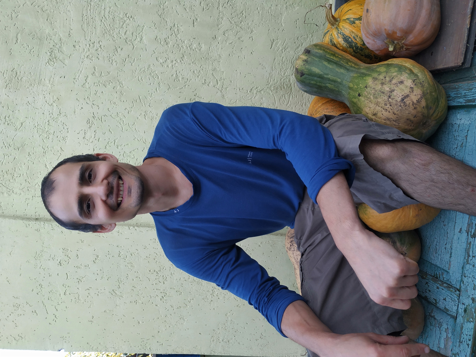 A man is sitting on a blue wooden bench with a light yellow wall in the background. The man is wearing a blue long-sleeved shirt and grey shorts. He has dark hair and a beard, and he is smiling at the camera. In front of him are several pumpkins of various sizes and colors, including a long green and yellow squash, a round orange pumpkin, and a round yellow and green striped pumpkin. The man's left arm is resting on his lap, and his right hand is on one of the pumpkins. The scene appears to be outdoors, possibly in a garden or yard.