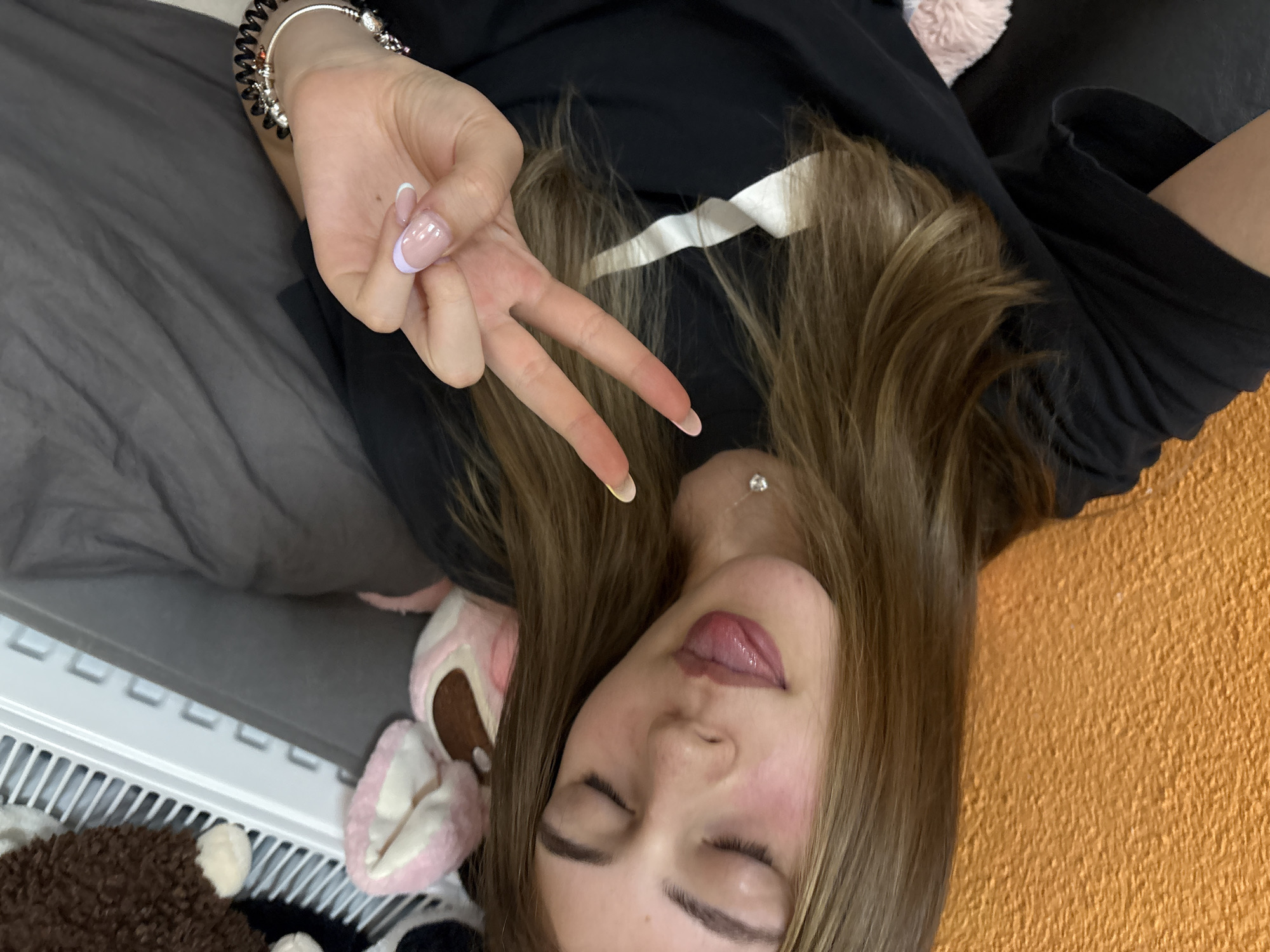 A young woman is lying down, possibly on a bed, in a room with an orange wall. She has long, light brown hair and is wearing a black shirt. Her eyes are closed, and her tongue is slightly sticking out. She is making a peace sign with her right hand, which has a silver bracelet on the wrist and neatly manicured nails with a light purple French tip. To her left are gray bedding and several stuffed animals, including one with pink ears. Part of a white radiator is visible in the lower left corner of the image. There is no clear indication that the image was taken in Panagyurishte, Bulgaria.