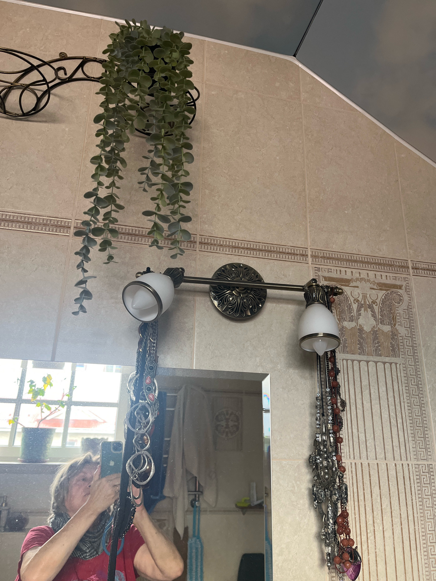 The image depicts a bathroom scene, likely in an apartment in Vladimirovka, Russia. The walls are tiled in a neutral beige tone with decorative borders. A faux hanging plant with numerous green leaves is mounted on a black metal bracket to the left. Below this, a double-headed vanity light fixture with white glass shades is attached to the wall. Hanging from the fixture are several strands of jewelry, including necklaces and bracelets.
In the foreground, a mirror reflects a person taking the photo with a smartphone. The person is wearing a red t-shirt and a scarf. Through the mirror, a window is visible, showing a potted plant on a windowsill. The overall impression is a functional, possibly dated, bathroom environment. The presence of a mirror suggests this is a vanity area.