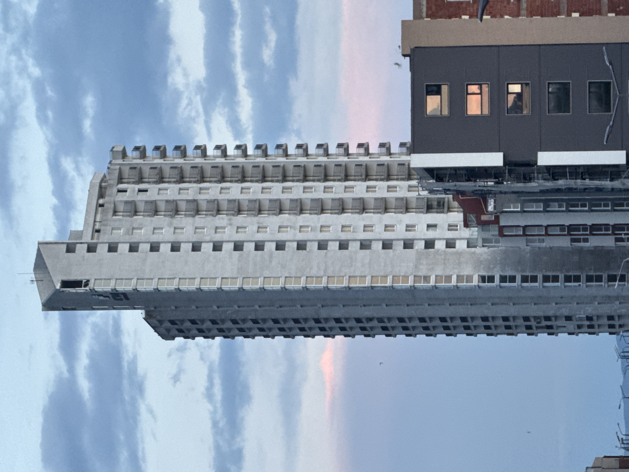 The image shows a large, white concrete building, likely an apartment complex, against a backdrop of a cloudy sky. The building has a distinctive tiered design, with numerous windows visible on each level. At the top of the building, there is a row of smaller, turret-like structures. To the right of the white building, a taller, more modern structure is visible. This building has a dark gray facade with a few windows reflecting sunlight. The sky in the background is a mix of blue and gray with pink accents. The image is rotated 90 degrees counter-clockwise.
Based on the architecture, the building is likely the Hotel Bulgaria in Burgas, Bulgaria, identifiable by its tiered concrete structure.