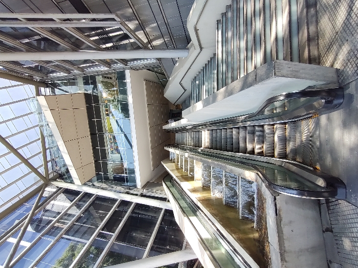 An indoor, overhead view captures escalators within a modern building. A pyramidal glass roof and a neutral-toned building section with glass windows contrast with the linear escalators. The escalators are constructed with steel, creating a functional, industrial design element. The photograph highlights the architectural integration of function and form within a bright, contemporary setting.