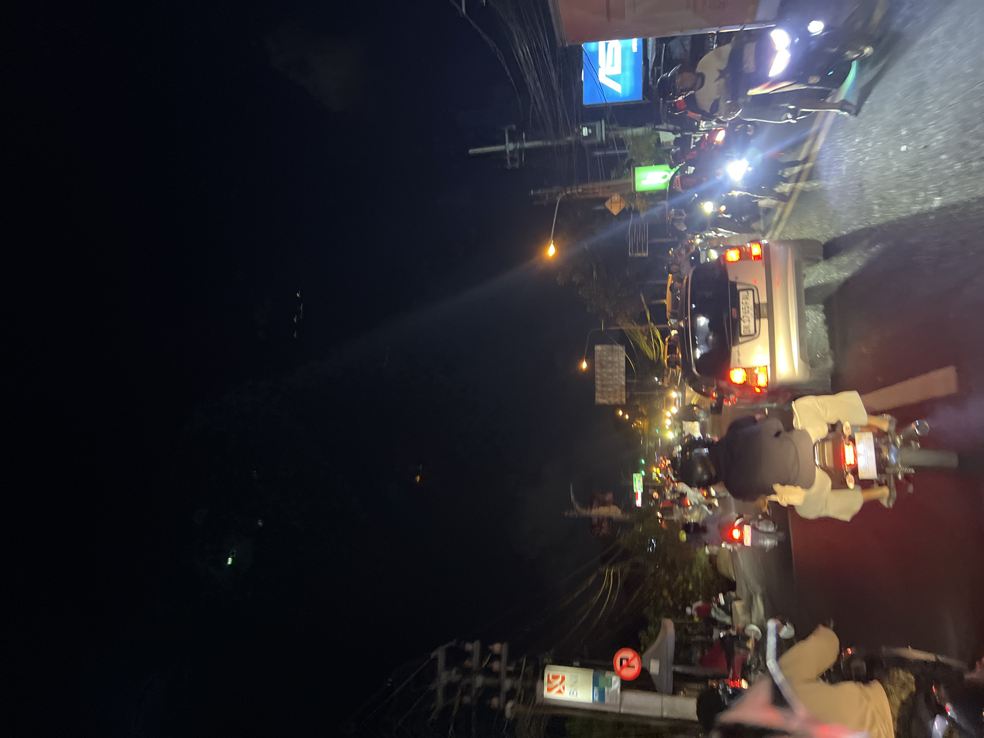 The image depicts a nighttime street scene in Denpasar, Indonesia, characterized by heavy traffic congestion. The perspective is from within a vehicle, looking forward at a road filled with motorcycles and cars. Several individuals are visible riding motorcycles, most wearing helmets. One motorcyclist in the foreground is wearing a dark jacket and light-colored pants, while another person in front of them wears a light-colored top and pants. Another individual on a motorcycle further down the road wears a light-colored shirt and a helmet. The road surface is asphalt, and the scene is illuminated by streetlights, vehicle headlights, and the glowing taillights of the vehicles. Buildings and signs are present on either side of the road. One sign on the left side features the letters "BNI" and a red circular sign with a diagonal line through a "P" symbol, indicating parking restrictions. A large blue sign with a white "ASUS" logo is visible on the right side. The sky is dark, indicating it is nighttime, with some diffused light sources visible in the distance. The overall impression is one of a busy urban environment at night.