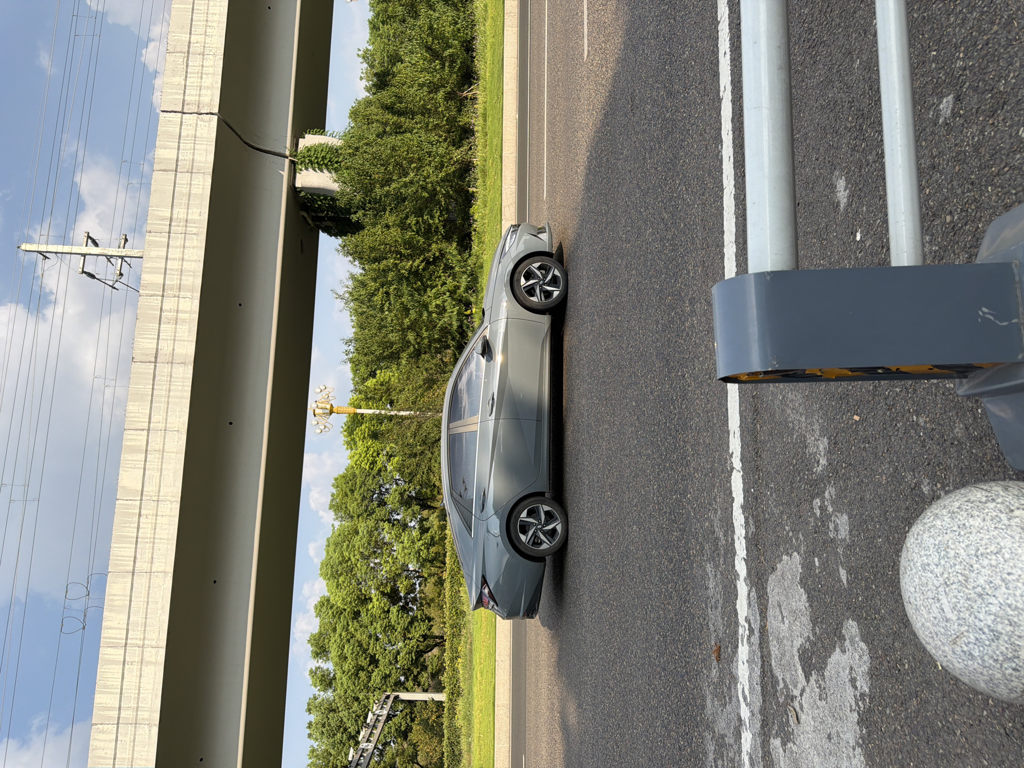 A gray sedan drives on an asphalt road beneath an elevated concrete structure. The car is positioned in the right half of the frame, traveling from left to right. The elevated structure spans the upper portion of the image, with power lines visible above it. Green trees and foliage form a dense line along the side of the road, creating a backdrop. A street light with a decorative top is visible amongst the trees on the right side. The road is marked with white lines. In the foreground, a gray protective barrier and a rounded granite bollard are present on the right side of the image. The scene appears to be in a suburban or semi-urban area with clear sky and scattered clouds.