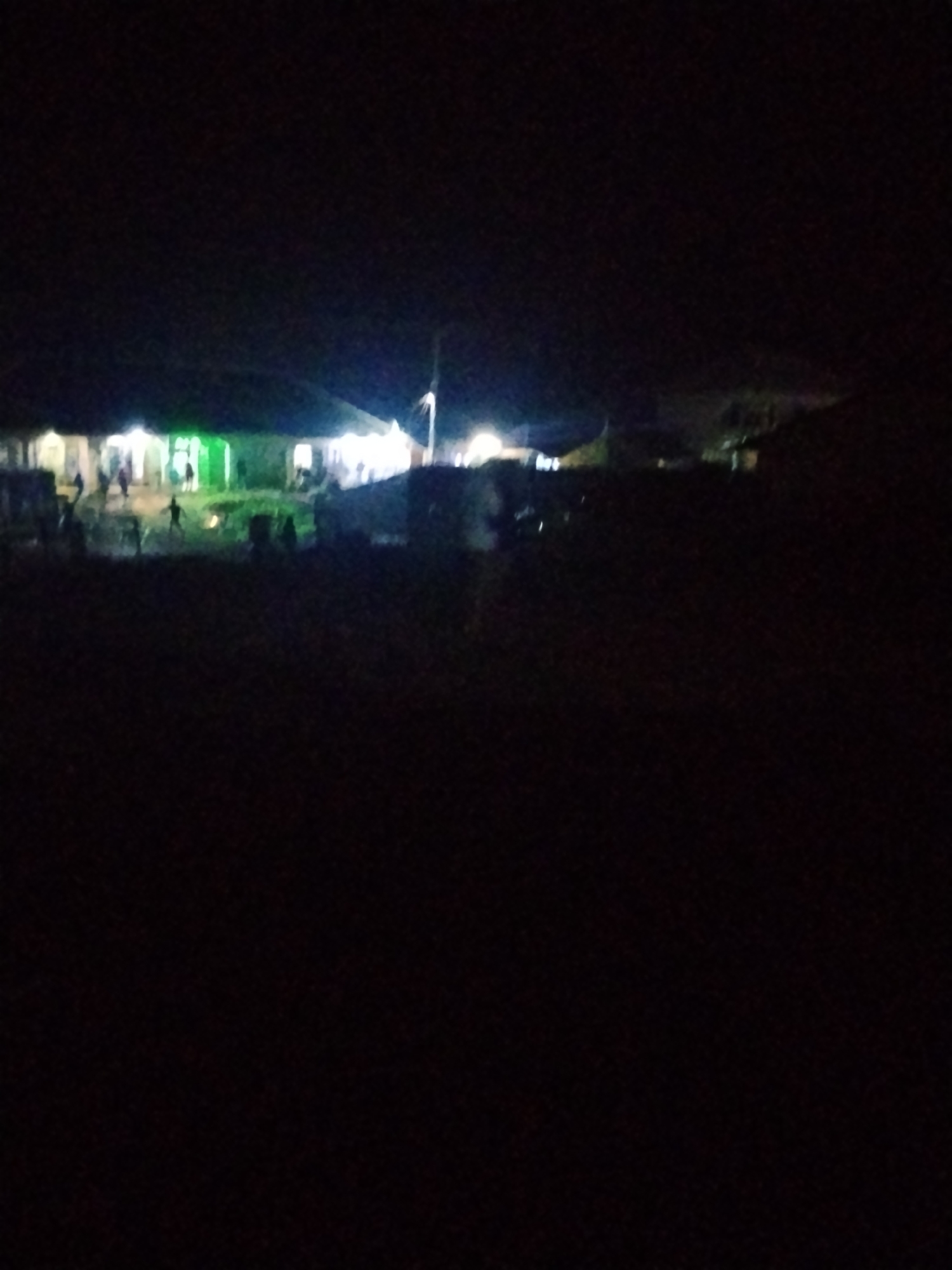 The image depicts a dimly lit scene at night, likely in Durumi, Nigeria. In the background, a building or complex of buildings is illuminated by artificial lights, casting a glow on the surroundings. Several figures, appearing to be people, are scattered around the illuminated area, some of whom are in motion. The foreground is predominantly dark, suggesting an outdoor environment with minimal ambient light. The overall impression is one of a gathering or activity taking place after dark in a somewhat rural or less developed urban setting.