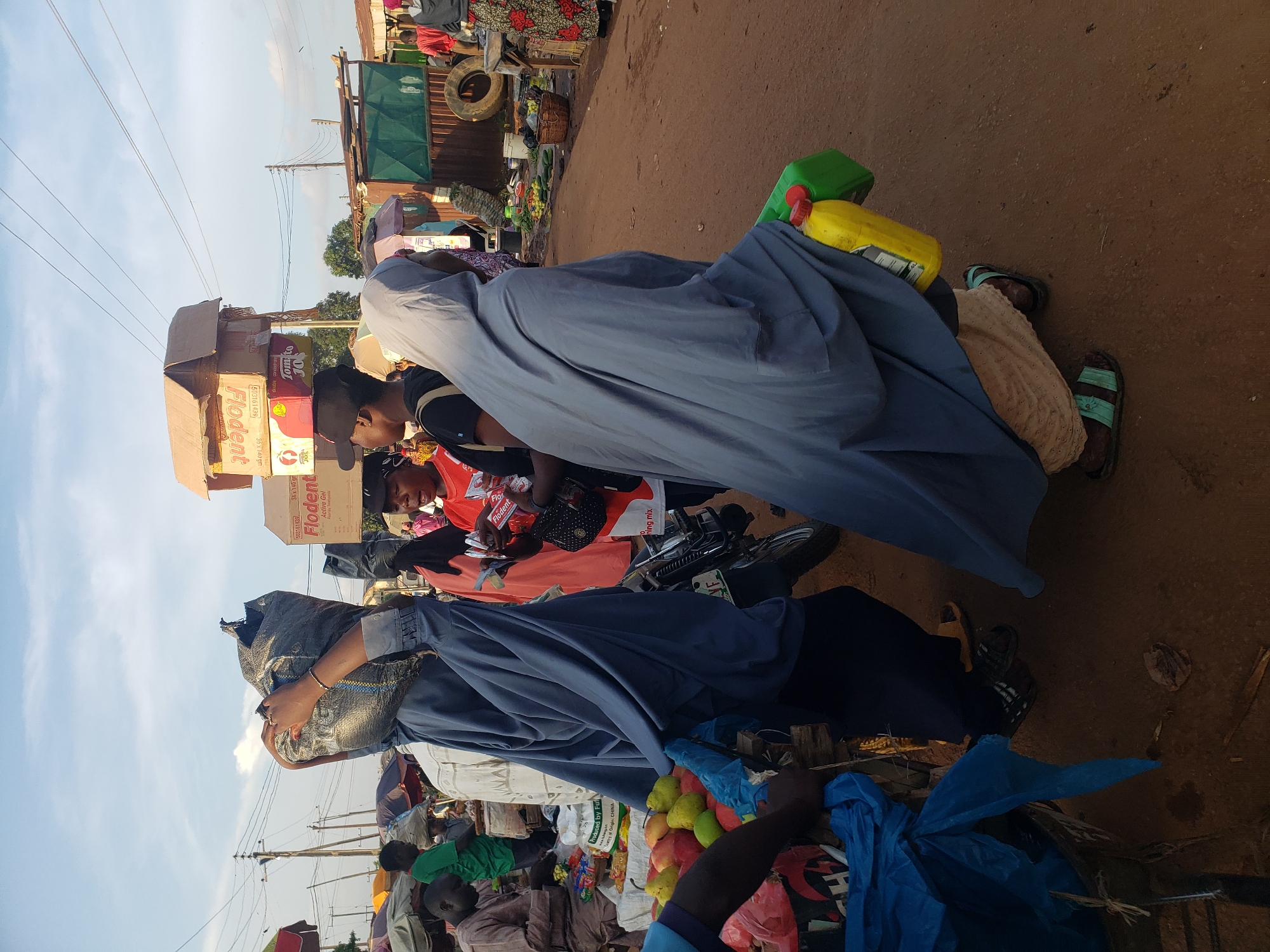 In Kakuri Gwari, Nigeria, a street market bustles with activity under a pale sky. A woman, her face obscured by a blue hijab, carries a large black bag on her head. Beside her, a person in a similar blue garment holds a yellow container and a green box. In the background, various market stalls are visible, with vendors and shoppers engaging in commerce. Produce, such as fruits, is displayed on a table, and boxes are stacked high. A motorcycle is parked near a stall. The ground is a dusty, reddish-brown dirt. The scene captures a slice of everyday life in a Nigerian market.