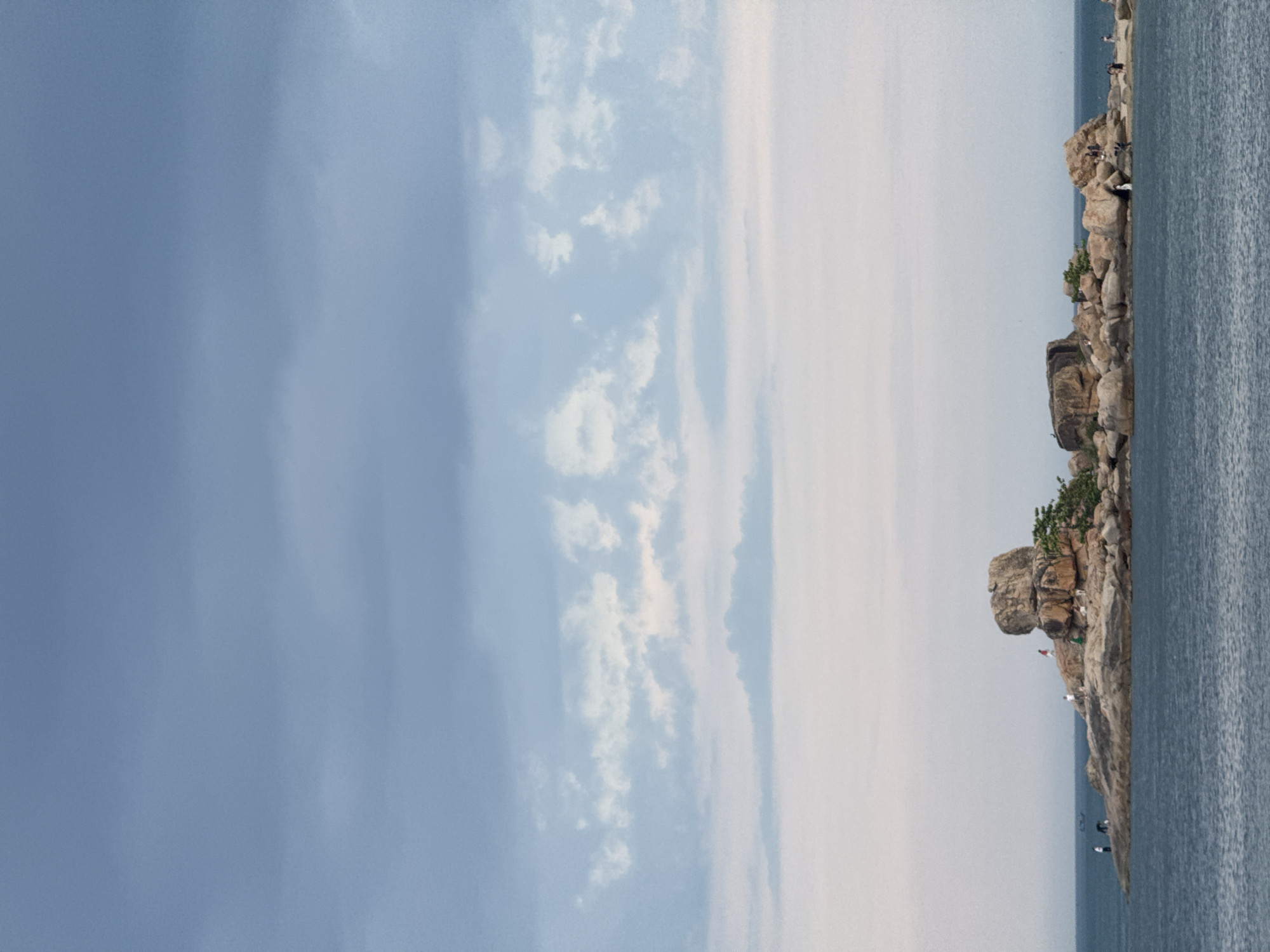The image depicts a coastal scene in Bac Nha Trang Ward, Vietnam. A rocky coastline is visible on the right side of the frame, with several large rock formations extending into the water. A small number of people are discernible on the rocks. The sky is predominantly blue, with scattered clouds visible towards the left side. The water appears calm with a rippled surface.