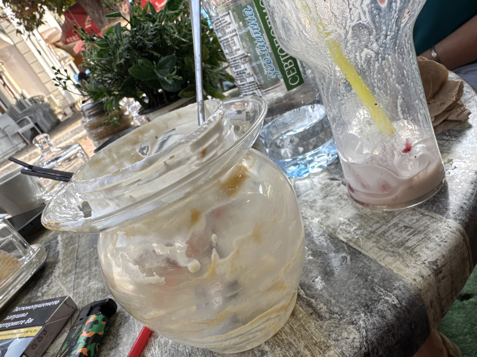 This is an outdoor, eye-level, close-up shot of a table at a cafe. The table is a dark, textured surface, possibly stone or concrete, and is cluttered with the remnants of a meal or drinks. In the foreground, a glass jar, likely used for a dessert or drink, is tilted and has a significant amount of residue inside. A metal spoon is also visible inside the jar. To the right, a clear glass cup, also with residue and ice, is present, with a yellow plastic straw sticking out. A bottle of water with a label is partially visible behind the jar. In the background, a green potted plant with delicate leaves is situated on the table. A dark cigarette pack and a camouflage-patterned lighter are on the left side of the frame. The overall impression is of a recently finished outdoor dining experience. The image does not contain any visual information that would allow for the identification of the city of Lovech, Bulgaria.