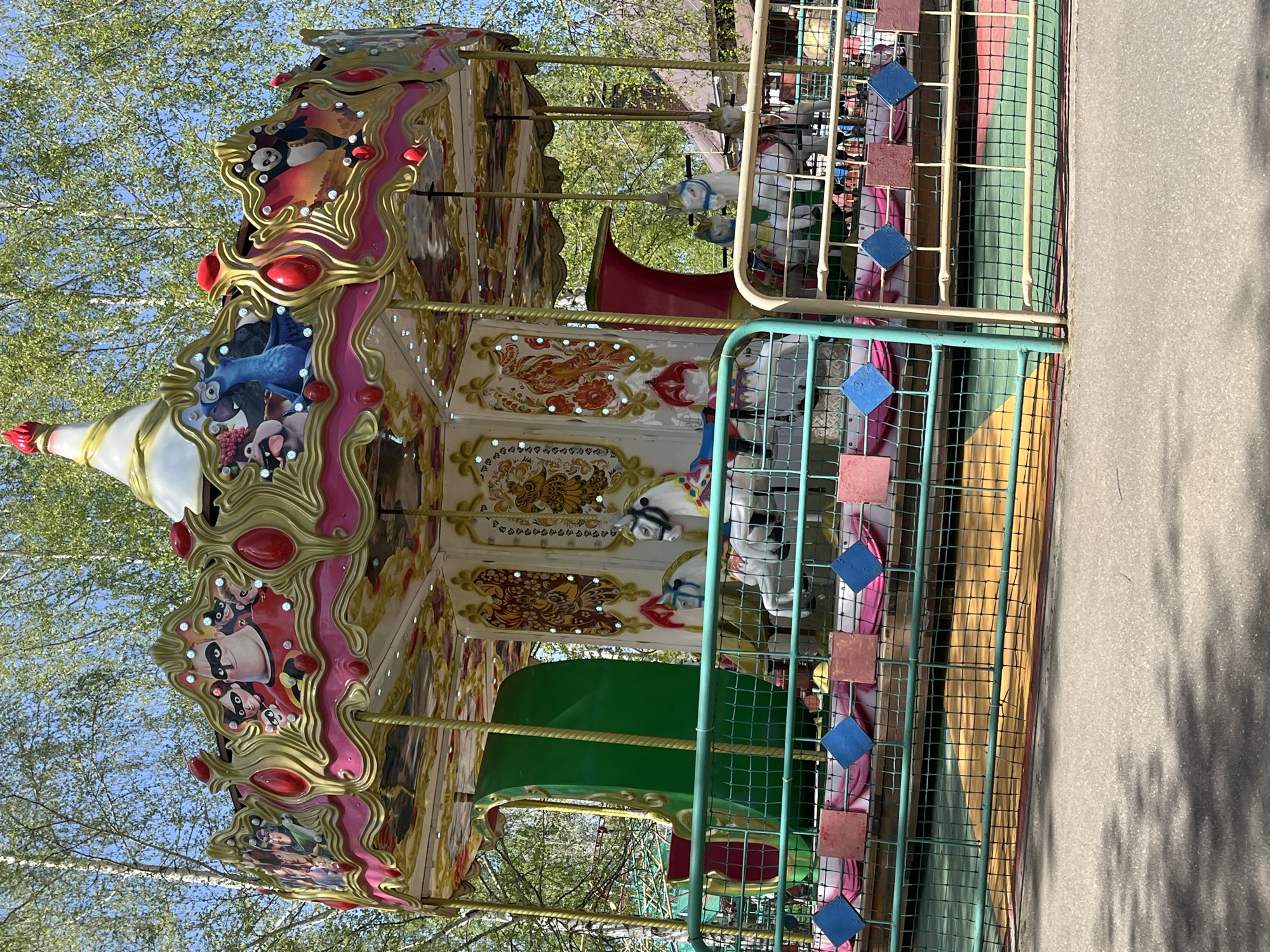 The image shows a carousel in Chaikovsky, Russia. The carousel is decorated with cartoon characters and has ornate detailing with gold trim, pink and red accents. It is surrounded by a green metal fence with blue diamond and red square accents. The ground is paved, and trees are visible in the background, suggesting the carousel is located in a park or outdoor recreational area.