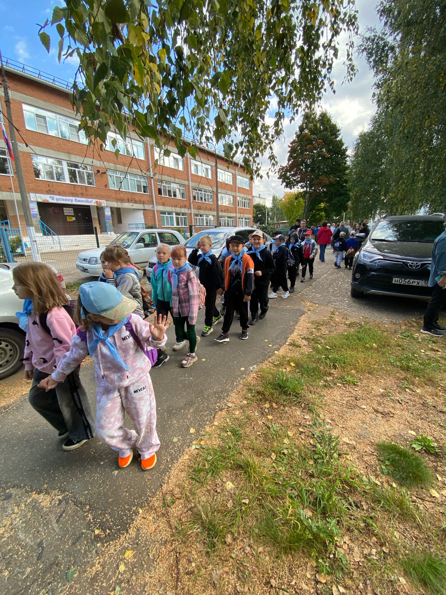 In Noviy, Russia, a group of children, some wearing matching blue neckerchiefs, are walking on a paved path next to a brick building with numerous windows. The building appears to be a school, as indicated by a sign above the entrance that reads "К успеху вместе!" (Together to success!). Several cars are parked along the side of the path, including a black SUV in the foreground. The path is lined with grass and fallen leaves, suggesting it is autumn. The children are mostly dressed in casual clothing, with backpacks on some of them. They are walking in a line, and some appear to be interacting with each other. The sky is partly cloudy.
