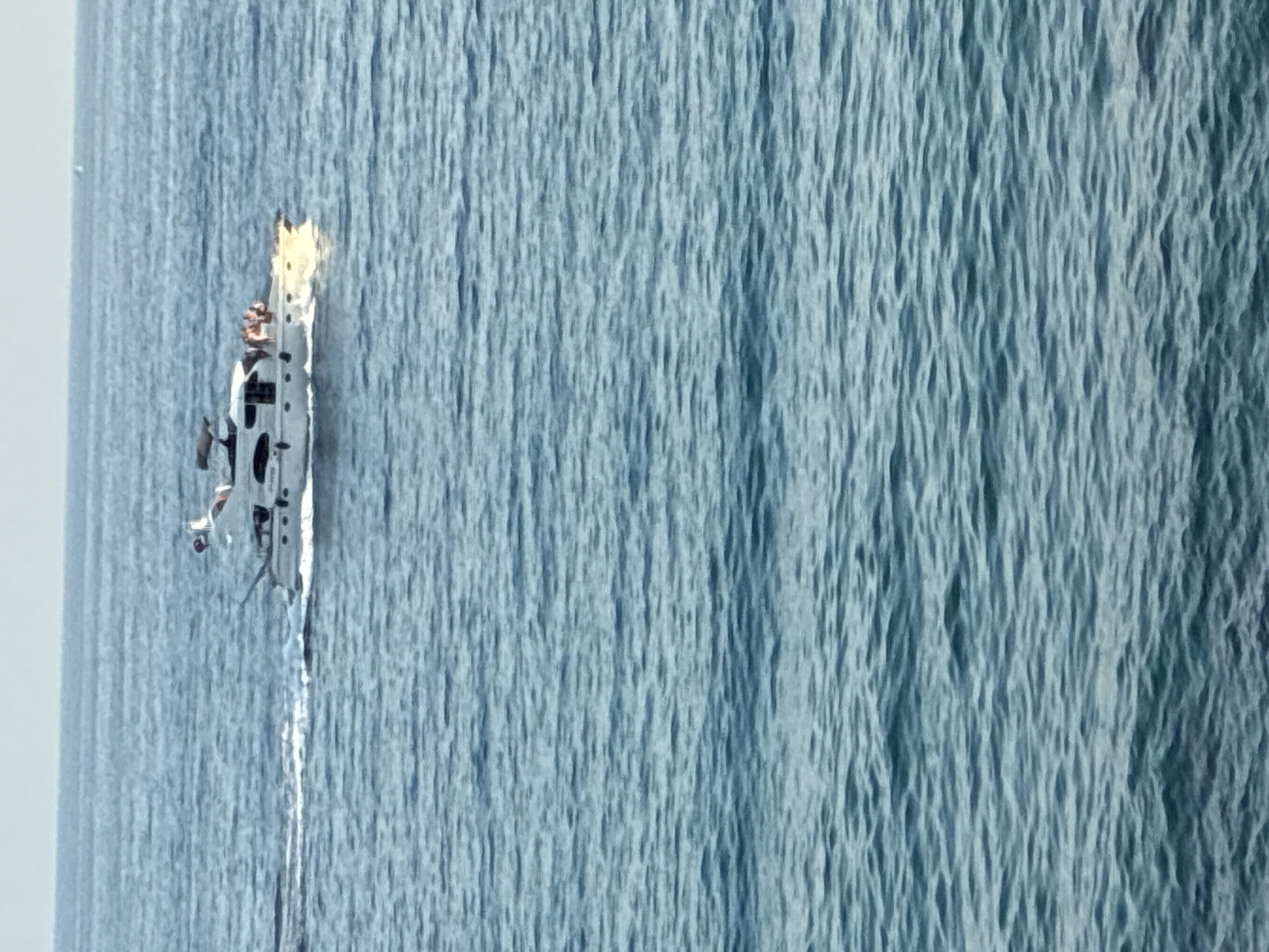 The image shows a small boat or a ship with a few people on board that appears to be on fire, with flames and smoke erupting from the top deck. The vessel is white with some black accents and has a somewhat dilapidated appearance, with parts of its structure seemingly damaged or torn. The boat is situated in the middle of a large body of water, which is a deep blue and appears to have a rippled texture, suggesting it is either the sea or a large lake. The background consists of more of the blue water, with a bright, slightly hazy sky visible on the left side of the image. The overall scene suggests an emergency or a dangerous situation occurring at sea. There is no visible text in the image. The lighting suggests it is daytime, but it's difficult to determine the exact time of day or the weather conditions beyond the presence of the water and sky. Given the location context of Alanya, Turkey, this could be the Mediterranean Sea. The activity depicted is clearly a fire on a boat with people aboard, indicating a potential distress event.