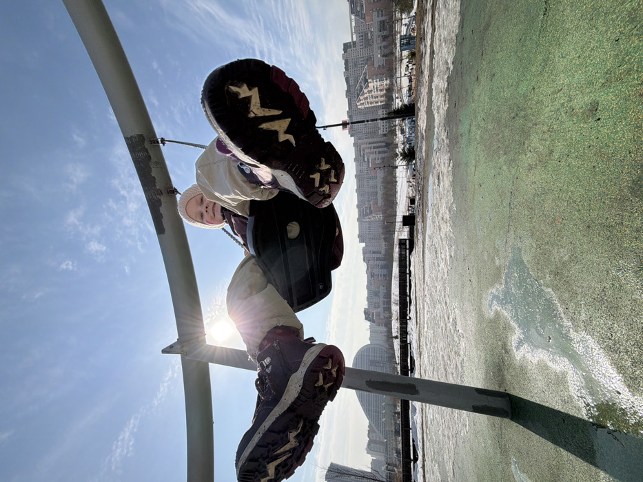 A low-angle photograph captures a young child seated in a swing within a playground, set against a bright, clear blue sky. The child, appearing to be a girl, wears a light pink knit beanie, light-colored winter trousers, and dark maroon/purple winter boots with prominent sole tread patterns (a white/yellow zigzag on the right boot, a star-like pattern on the left). Her legs are extended upwards and forwards, and she looks towards the camera with a slight smile. The swing seat is dark-colored. The sun is positioned to the left, causing lens flare and backlighting.
The swing apparatus consists of a large, curved, light gray or silver metallic bar supported by vertical posts, one visible on the left side of the frame. The curved bar exhibits areas of discoloration and wear. Below the swing, the ground is covered in a green, textured safety surface, partially obscured by patches of melting snow and ice. A long, dark shadow of a support post is cast on this surface.
In the mid-ground, beyond the playground, lies a partially snow-covered park area with sparse deciduous trees and a small coniferous tree. In the far background, a dense cityscape is visible under a distant haze. Key architectural features include a tall, slender high-rise building on the far left, a distinctive modern building with a large, curved, wave-like roof structure in the center-left, and a continuous expanse of multi-story residential buildings featuring terraced or stepped facades. This urban landscape is located in the city of Khimki, Moscow Oblast, Russia, characteristic of modern developments in areas such as the Levoberezhny district, which features such contemporary architecture including structures near the Khimki Arena. The overall scene is brightly lit, indicative of a sunny day with the sun relatively low in the sky, casting long shadows.