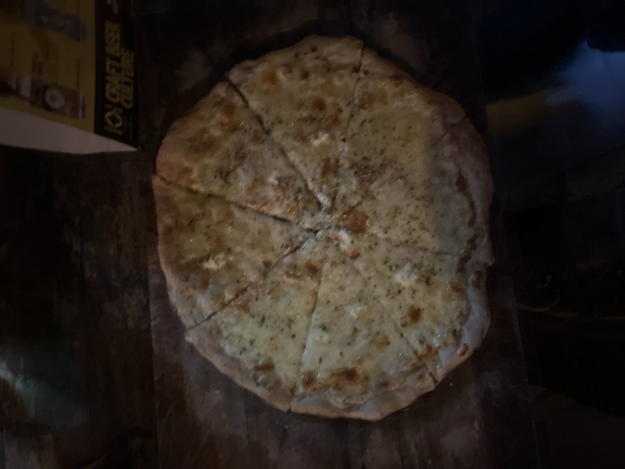 A round pizza, generously covered with melted cheese and sprinkled with dried herbs and black pepper, is the central subject of the image. The pizza appears to be a white or cheese pizza and is neatly cut into eight slices. It rests on a dark, rustic wooden surface, likely a pizza board or table, which shows visible grain and texture.
The overall scene is dimly lit with warm light, suggesting an evening setting in a casual indoor environment, such as a restaurant or bar. The low light creates prominent shadows around the pizza and in the background.
To the upper left of the pizza, a portion of a sign or menu is visible. The legible text on this dark-colored sign, in yellow or gold lettering, reads "ION CRAFT BEER CULTURE." This detail provides a cue about the establishment's theme. No people are visible in the frame, keeping the focus entirely on the pizza and its immediate surroundings. The image captures a ready-to-eat meal in a cozy, subdued atmosphere.