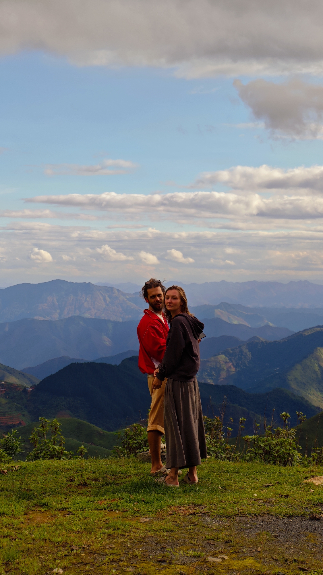 A man and a woman are standing on a grassy, elevated area overlooking a vast mountainous landscape in Xín Vàng, Vietnam. The man, positioned on the left, has shoulder-length brown hair and a beard, with sunglasses resting on his head. He wears a red jacket over a white shirt and light brown shorts, with light-colored sandals visible on his feet. His right arm is around the woman. The woman, standing to his right, has long, light brown hair and is wearing a dark grey hooded sweatshirt, a long dark grey skirt, and light-colored sandals. Both individuals are looking directly at the camera, the man with a slight smile and the woman with a neutral expression. The background features numerous layers of mountains receding into the distance, appearing progressively bluer due to atmospheric haze. Some distant slopes show signs of terraced cultivation. Above the mountains, the sky is predominantly blue with scattered white and grey clouds. The foreground consists of green grass interspersed with patches of bare earth and small rocks.