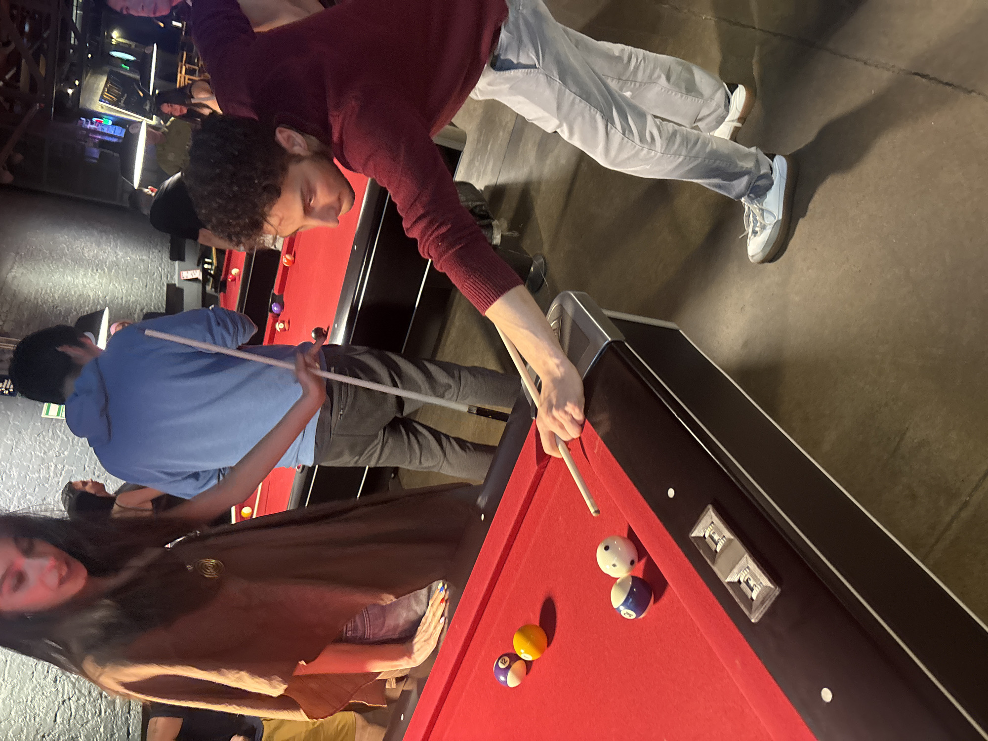 A slightly rotated medium indoor shot features people around a pool table, with a focus on a man in a burgundy sweater playing a game. The pool table is covered in red felt, and several balls are visible. The man playing is leaning over the table, cue in hand, with a focused expression. A person in a blue shirt stands to his left, holding another cue. Further to the left, a woman with long dark hair in a brown jacket stands near the table. Other people are present in the background, some near another pool table. The location is possibly a bar or recreational establishment. Given the background elements and the presence of a pool table, the location could be a pool hall or a bar with recreational facilities. The image doesn't provide explicit cues to confirm it's in Mexico City, Mexico.