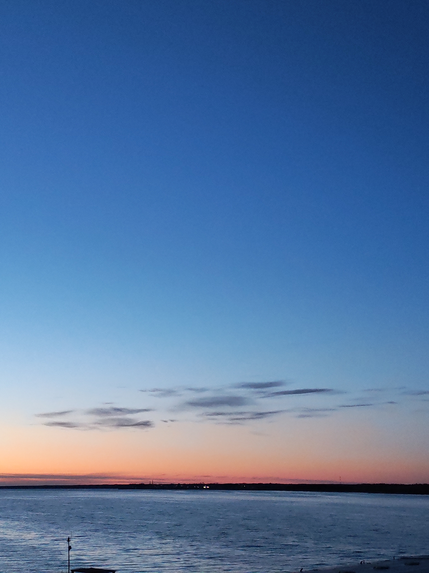 The image captures a serene twilight landscape, likely at either sunrise or sunset, dominated by a broad expanse of sky and a large body of water.
The upper two-thirds of the frame are occupied by the sky, which exhibits a clear gradient of blue hues. At the very top, the sky is a deep, rich royal blue, gradually lightening to a softer, clearer blue as it approaches the horizon. A band of sparse, elongated, dark grey clouds is visible horizontally across the middle of the sky. Below these clouds, the sky transitions into a delicate blend of soft orange and pink, indicating the fading or rising sun.
A thin, dark silhouette of land, possibly a distant shoreline with trees, stretches across the horizon, separating the sky from the water. Faint points of light, suggesting a distant settlement or industrial structures, are discernible along this dark landmass.
The lower third of the image is filled by a calm body of water, which appears as a muted blue-grey with subtle ripples on its surface, reflecting the dim light of the sky. In the bottom left foreground, a dark, partially submerged structure or pole rises from the water or shoreline, with some faint details suggesting attachments. The very bottom right shows indistinct, dark shapes on what might be a sandy or pebbly bank.
The overall impression is one of tranquility and vastness, with the contrasting colors of the sky being the most prominent feature. No people or discernible activities are taking place, and there is no visible text.