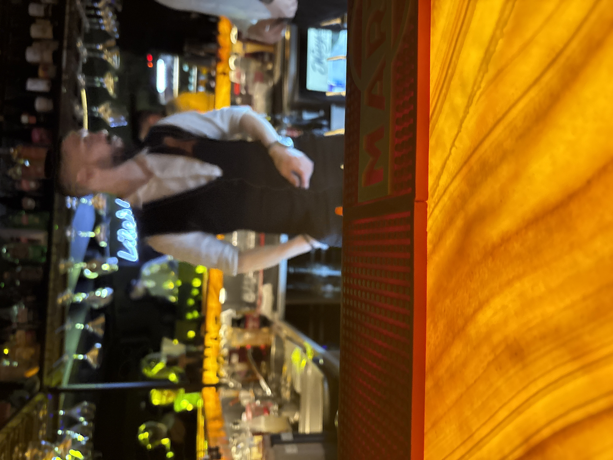 Here's a detailed description of the image:
The image appears to be taken in a bar, potentially located in Perm, Russia, based on the "MAX" signage. A male bartender, wearing a white shirt and a black vest, is positioned behind the bar. A row of liquor bottles and glasses lines the back of the bar. In front of the bartender is a workstation likely used for preparing drinks. The environment is dimly lit with multiple sources of lighting that create a warm and inviting atmosphere. The bar has a counter with amber panels on one side.