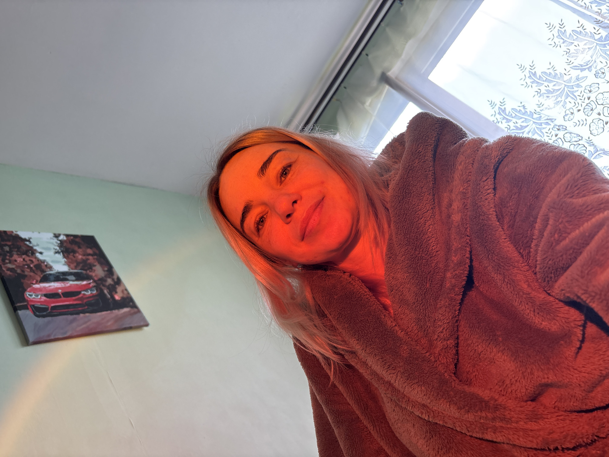 The image captures a close-up, low-angle shot of a woman indoors. She has blonde hair and fair skin, and is looking directly at the camera with a subtle, pleasant smile. Her face is bathed in a distinct, warm red-orange light, while the rest of the scene has more natural ambient lighting. She is wearing a soft, brown, hooded robe or garment that appears fluffy in texture.
In the background, to the left, a framed painting or canvas is visible on a light green wall. The painting depicts a red sports car, possibly a BMW M4, on a road flanked by trees with reddish foliage, suggesting an autumnal scene. Above the woman's shoulder and to the right, a large window with white framing is visible, allowing bright natural light into the room. Sheer white curtains with a delicate blue or gray floral and botanical pattern are drawn across the window. The ceiling is a plain light gray or white.
The activity taking place is a selfie or a portrait shot. The warm red lighting on the woman's face gives the image a cozy or intimate atmosphere. No specific time of day can be definitively determined, though the bright window light suggests daytime. There is no visible text in the image.
Location context: Unknown, Unknown