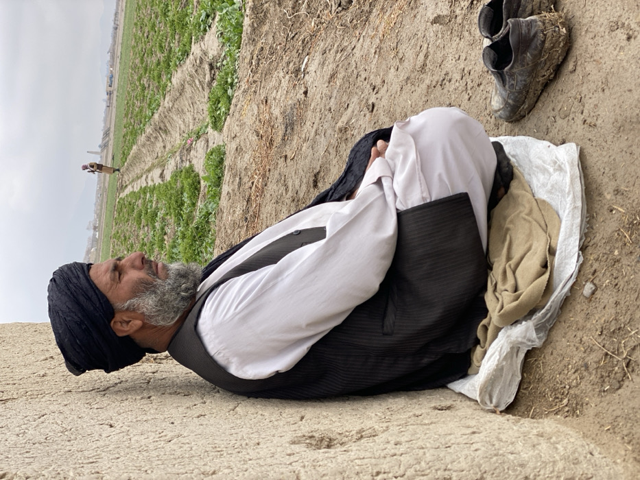 An elderly man with a turban and a gray beard rests on a makeshift bed of cloth and a crumpled plastic bag beside a muddy field. He wears a white shirt and a black vest. Dirty boots sit nearby. In the background, a distant figure tends the rows of crops under a cloudy sky. The scene suggests a hardscrabble rural life, with the man taking a break from agricultural labor.