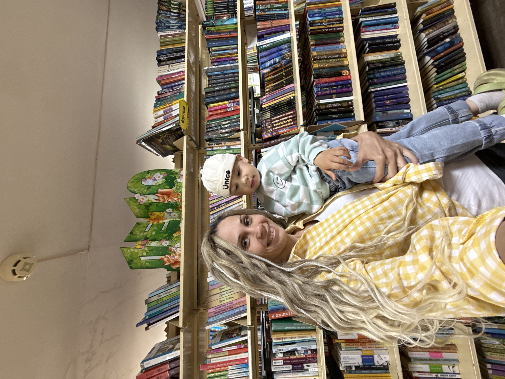 The image captures a heartwarming moment between a woman and a young child in what appears to be a home library or a cozy bookstore in Rusanovo, Russia. The woman, with long, wavy blonde hair featuring some braids, wears a cheerful yellow and white checkered shirt and smiles warmly at the camera. She is holding a small child, likely a boy, who is nestled across her lap, dressed in a light green and white checkered long-sleeve top, blue jeans, and a white cap bearing the text "Unice." The child also looks towards the camera with a gentle expression.
The background is dominated by numerous wooden bookshelves packed tightly with a vast collection of books, most with Cyrillic titles. The books are stacked vertically and horizontally, indicating a well-loved and extensively used collection. Among the visible titles are "ДЕТИ СОЛНЦЕВЫХ" (Children of the Sun), "ЖИВАЯ КАРТА" (Living Map), "МАЛЕНЬКИЙ ПОДАРОК" (Small Gift), "ТРИ ЛЕГЕНДЫ" (Three Legends), "ЧЕТЫРЕ ЖЕЛАНИЯ" (Four Wishes), "МОЯ ТЕТУШКА-ВЕДЬМА" (My Aunt-Witch), and multiple instances of "СТРАНЫ МЕЧТЫ" (Lands of Dreams). A yellow tag on one shelf clearly labels a section as "Фэнтези" (Fantasy). Decorative green cutouts featuring cartoon foxes and flowers are also visible on the shelves. A white smoke detector is mounted on the light-colored wall, which shows some minor streaks or discoloration, suggesting a lived-in space. The overall atmosphere conveys a sense of comfort, happiness, and a love for reading.