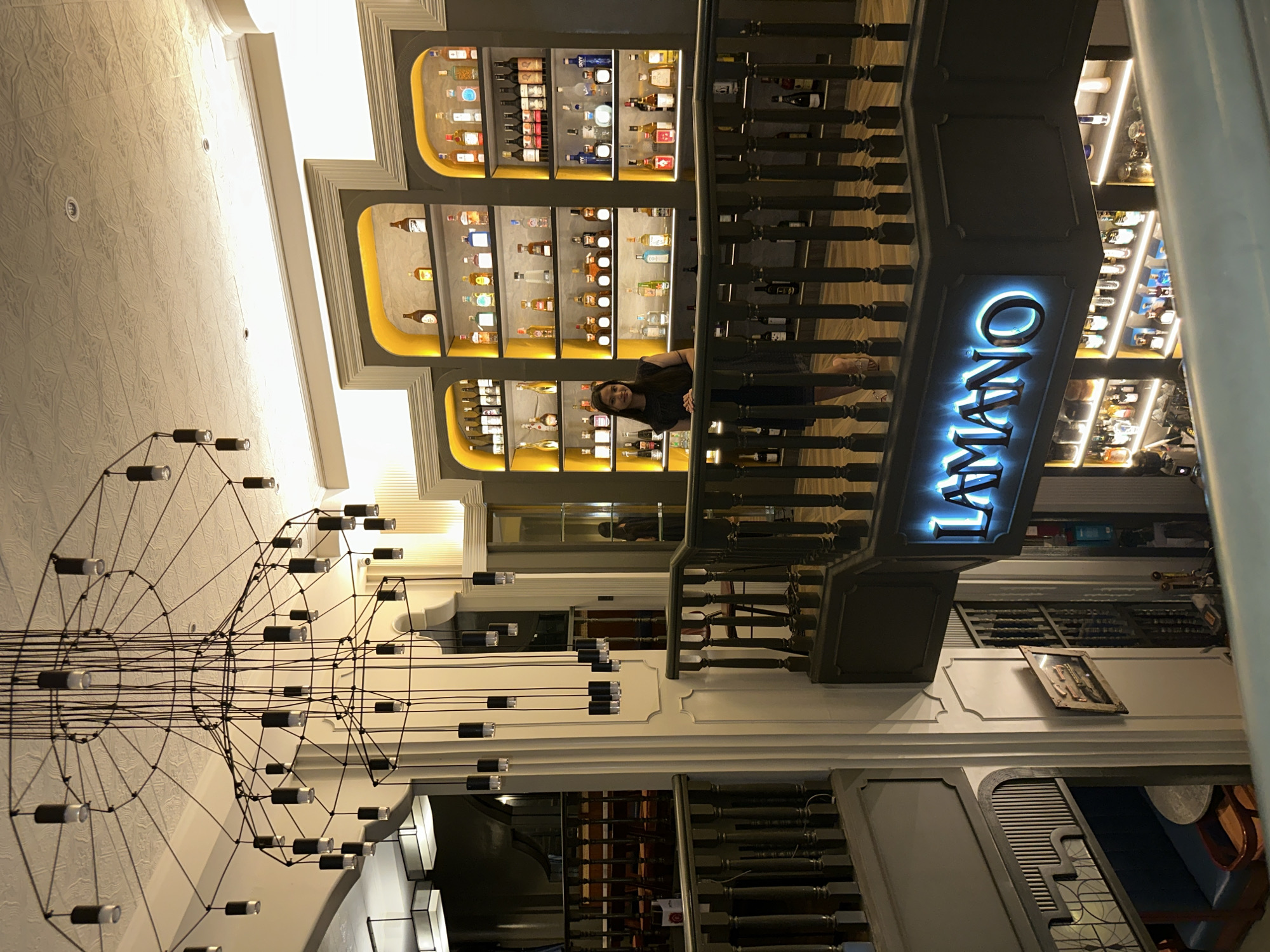 The image shows the interior of a well-lit establishment, likely a restaurant or bar. A woman in a dark dress is standing on a balcony overlooking the lower level. Behind her are illuminated shelves stocked with numerous bottles, suggesting a bar or liquor display. The architecture features a dramatic, multi-tiered chandelier hanging from the ceiling. The overall aesthetic is modern and sophisticated. The sign "LAMANO" is prominently displayed in neon lights on the balcony railing. The location is identified as Davao City, Philippines.