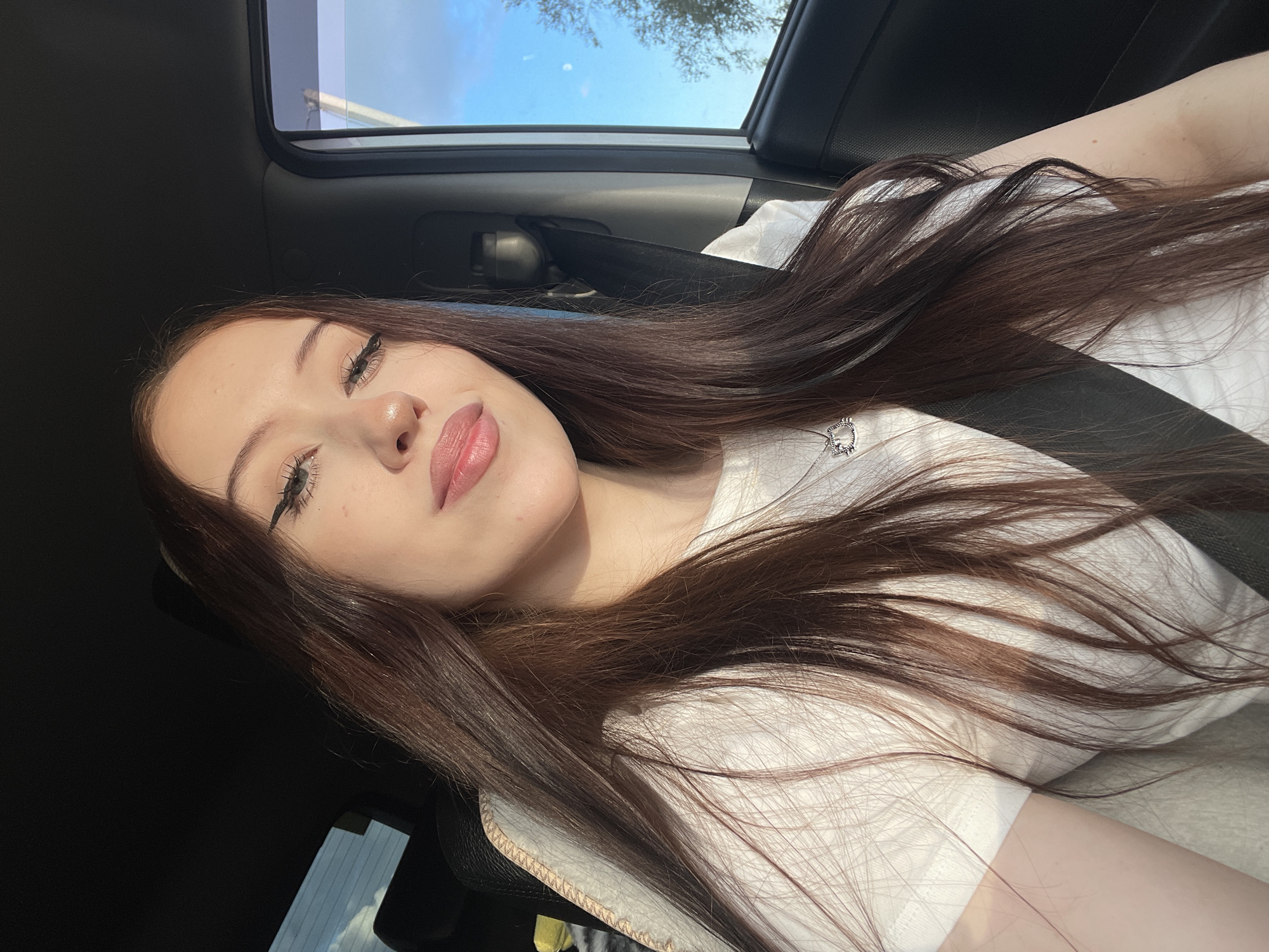 The image is a selfie taken from a low angle inside a car. The main subject is a young woman with long, dark brown hair, which is spread across her shoulders and chest. She is wearing a white t-shirt with a small design on the left side of her chest. Her face is partially visible, with her eyes closed and her lips slightly parted, suggesting a relaxed or possibly sleepy expression. She has winged eyeliner and pink lipstick. A black seatbelt is visible across her chest.
The setting appears to be daytime, with sunlight filtering into the car from the open sunroof and the side window. The sky visible through the sunroof is blue with some clouds, and trees can be seen in the distance, indicating an outdoor location, likely a rural or suburban area given the context of Gavrilova, Russia. The interior of the car is visible, including dark upholstery and the roof lining.
The overall mood of the image is casual and personal, typical of a selfie. The lighting suggests it is either late afternoon or early morning, given the warm, directional sunlight. There is no visible text in the image.