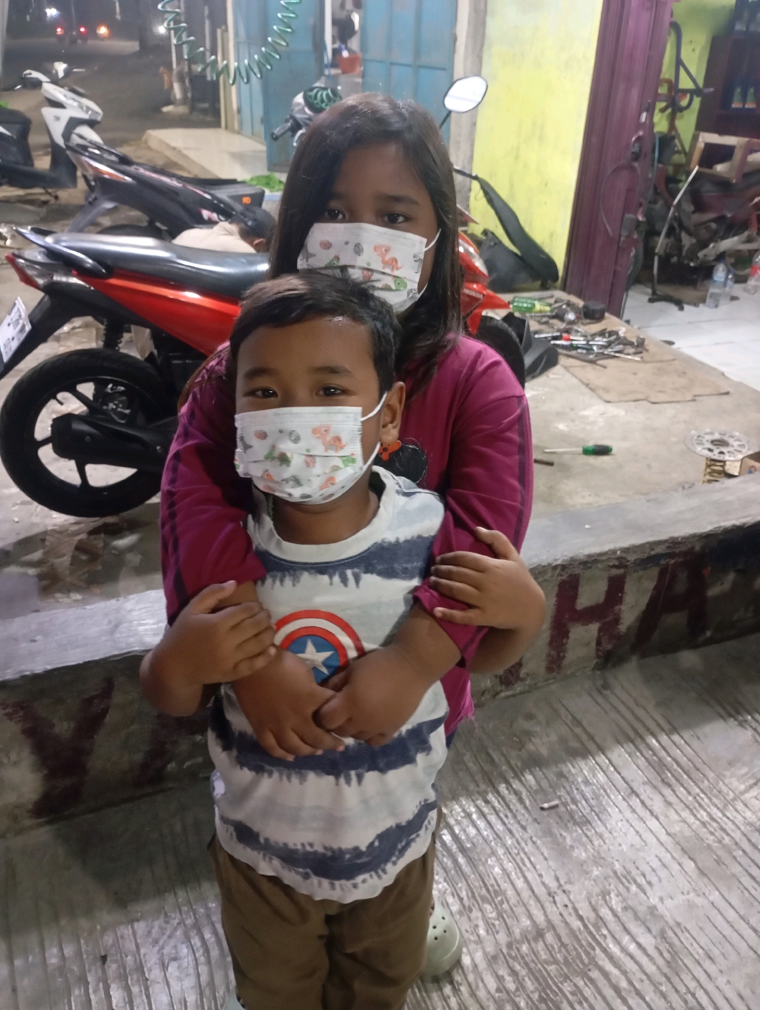 In an outdoor urban environment, likely a workshop or garage in Babelan, Indonesia, two children, a girl and a younger boy, are positioned in the foreground. Both children are wearing face masks. The girl, with dark hair, is standing behind the boy and embracing him. She is wearing a dark pink long-sleeved top. The boy, facing the camera, is wearing a striped t-shirt with a Captain America shield emblem on the chest and brown pants. He is also wearing a white face mask with a colorful cartoon pattern.
Behind the children, a red motorcycle with black accents is parked. The background includes various elements indicative of a workshop: tools are scattered on a concrete surface, including screwdrivers and what appears to be a brake disc. A yellow wall and a dark purple door or panel are visible in the background, along with other motorcycles and debris, suggesting a cluttered but functional space. The ground is a textured concrete surface. The lighting appears to be artificial and somewhat dim, possibly from overhead lights or shop lighting. The overall scene suggests a candid moment captured in a working area.