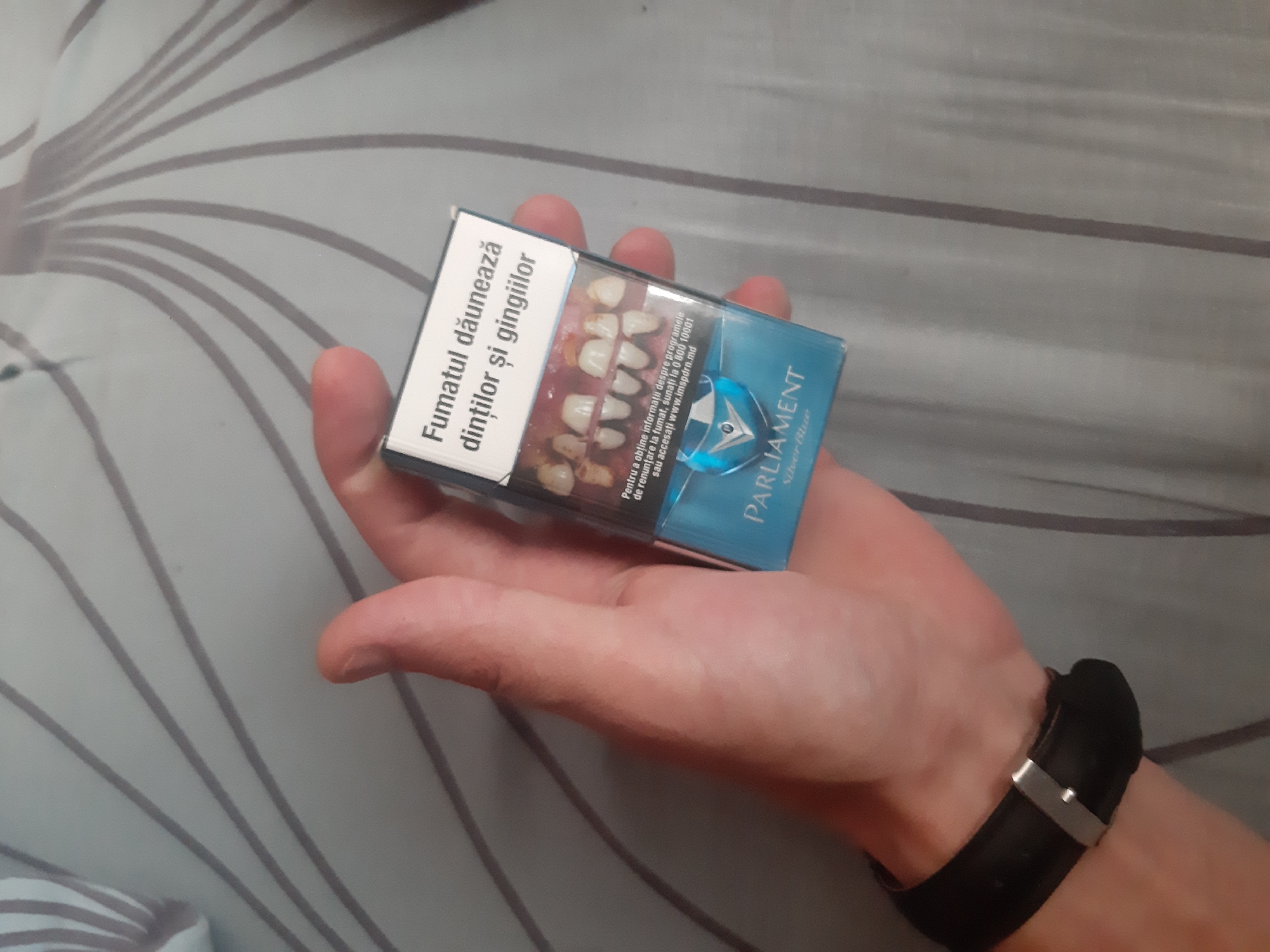 A person's hand is holding a pack of Parliament Silver Blue cigarettes. The pack displays a warning in Romanian about smoking damaging teeth and gums, accompanied by a graphic image of diseased teeth. The background is a gray striped fabric, possibly a bedsheet or sofa. The scene is set in Cricova, Moldova. The person is wearing a black watch on their wrist.