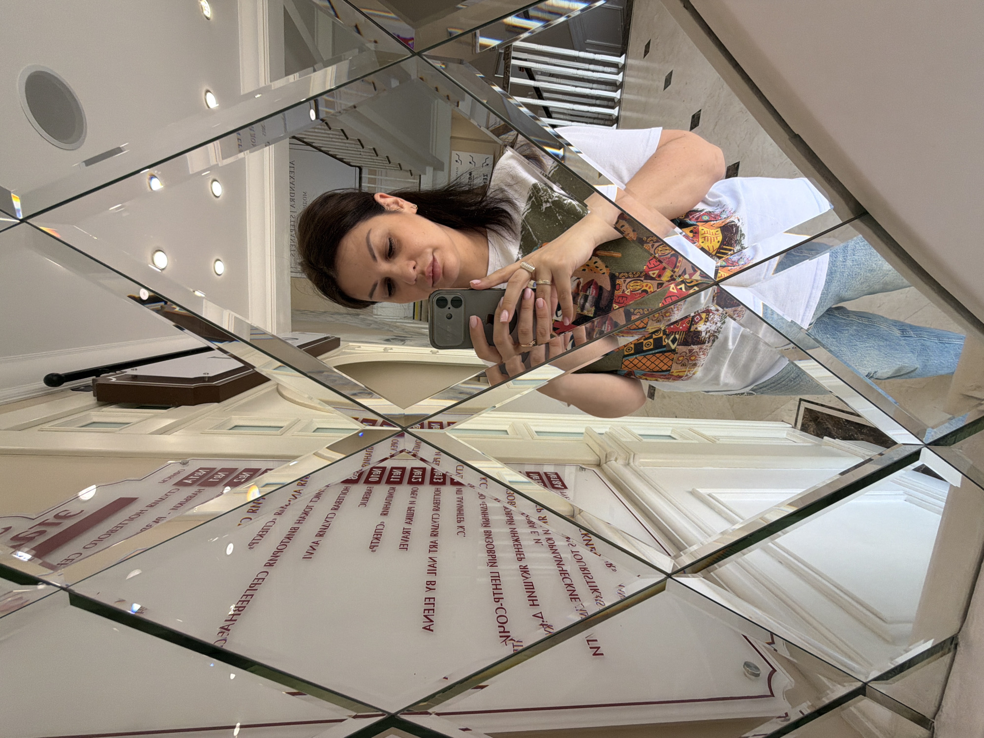 A woman is depicted taking a selfie in a multi-paneled, diamond-patterned mirror, which fragments her reflection and the background into various geometric shapes. The subject is a mid-adult female with dark hair, wearing a white t-shirt featuring a colorful, abstract graphic design and light blue jeans. She holds a dark-colored smartphone in her right hand, looking down at the screen. Multiple rings are visible on her fingers, and her fingernails are painted a light color.
Reflected in the mirror panels, directly behind the woman, is a large white wall displaying multiple lines of reversed red Cyrillic text. Several words are legible, including "ИЧОС" (reversed for "Sochi"), confirming the location as Sochi, Russia. Other decipherable reversed text includes names such as "ЭЛЕНА" (Elena), "АННА" (Anna), "АЛЕКСАНДРА" (Alexandra), and "РУСЛАН" (Ruslan), alongside terms like "ОБЩЕСТВО" (Society) and "ДЕЛОВОЕ" (Business), suggesting a business directory, list of associates, or event participants. Numbered entries such as "№1", "№10", and "№13" are also present.
To the right of the woman's reflection, a white staircase with a dark handrail ascends, indicating a multi-story interior space. The ceiling, visible in upper mirror panels, is white and features several recessed circular lights. The floor, reflected in the lower panels, is composed of light-colored tiles with darker square inlays. The environment appears to be an elegant and well-lit interior, likely a lobby or hallway of a commercial or upscale residential building in Sochi, Russia.