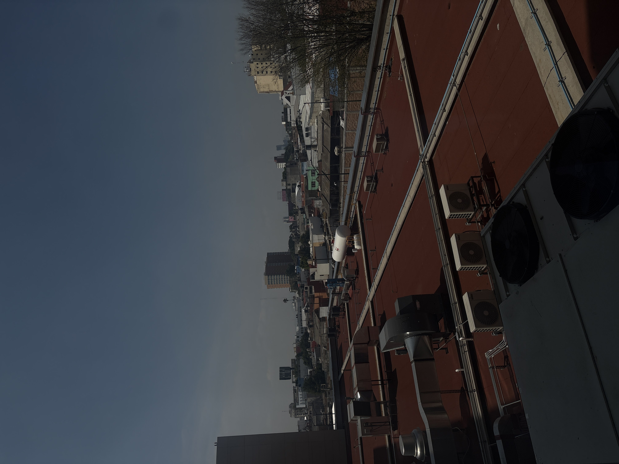 The image displays an elevated rooftop perspective overlooking an urban environment, identified as Puebla, Mexico. The foreground is dominated by a wide, flat, reddish-brown rooftop, densely covered with various industrial and HVAC equipment. This includes multiple air conditioning units, some with prominent circular fans, silver and dark gray ventilation ducts, and an intricate network of pipes and electrical conduits. A large, white cylindrical tank, likely a water heater or gas tank, and a smaller silver tank are also visible on this roof.
Beyond the immediate rooftop, the scene transitions to a sprawling cityscape under a vast, clear blue-gray sky. The urban landscape features numerous buildings of diverse heights and architectural styles, stretching into the hazy distance. A notably tall, dark building is visible in the mid-ground. Bare-branched trees are interspersed throughout the urban fabric, suggesting either a winter season or a dry period.
The lighting indicates daytime, with soft light and shadows suggesting either the morning or late afternoon. The sky is free of clouds, though a slight atmospheric haze is present over the distant buildings. No people or active events are depicted; the scene is a static observation of the urban landscape. No visible text can be discerned on any objects or structures.