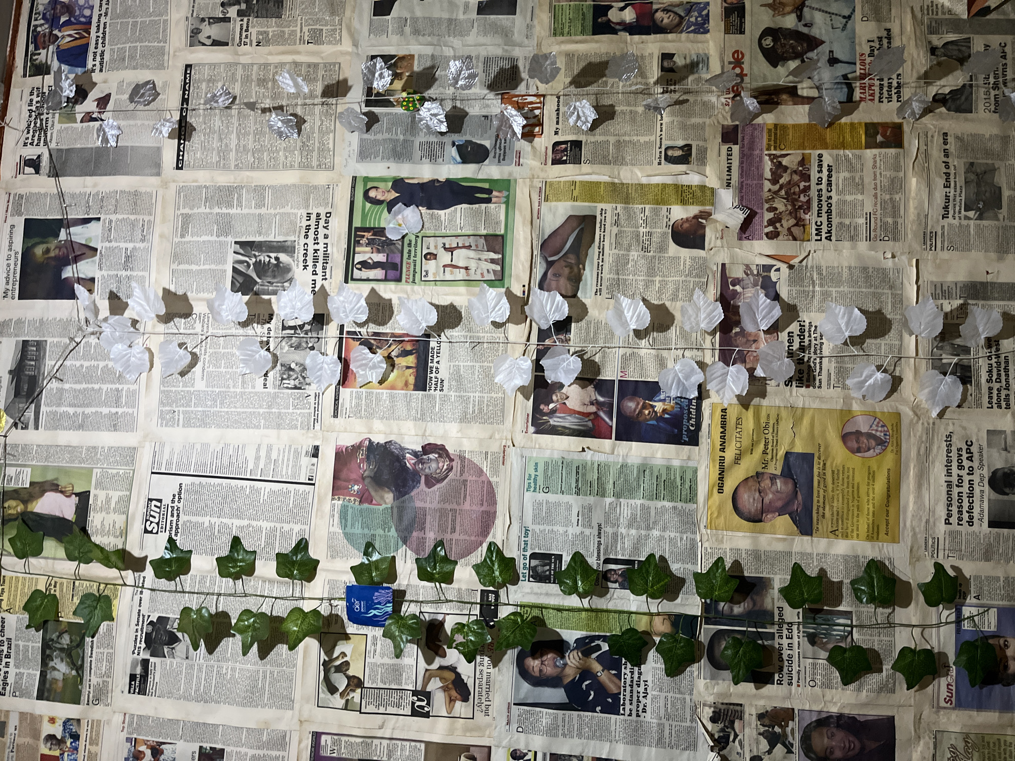 This is a close-up, eye-level, indoor shot of a wall covered in newspapers. The newspapers are layered and overlap, creating a textured collage. Various articles and photographs are visible, including portraits of people, sports action, and headlines. A string of artificial ivy with green leaves is draped vertically across the center of the image, with white paper leaves also attached to strings hanging down. There are also some blue and silver items affixed to the wall among the newspapers. The lighting appears to be artificial and casts shadows. The overall impression is of a densely covered wall, likely a decorative or informational display. The image does not provide any information about the city of Rafin Par, Nigeria.