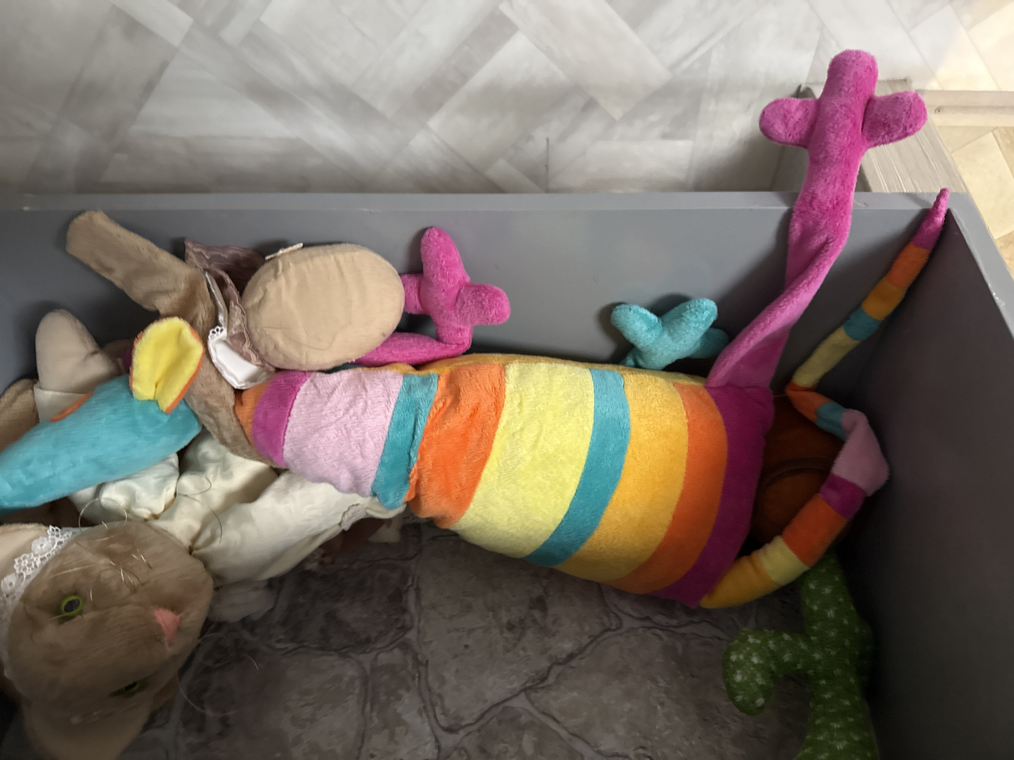 The image displays an interior setting featuring multiple stuffed animals positioned on a grey shelf. Dominantly, a large, horizontally oriented stuffed animal, resembling a chameleon or lizard, occupies the central space. Its body is segmentally striped in distinct colors: purple, orange, yellow, turquoise, orange, yellow, and pink from left to right. It has multiple limbs, with two turquoise limbs visible to its left and two pink limbs to its right, one of which extends below the shelf. Its tail, exhibiting similar striped coloration, also descends from the shelf.
To the upper left, a green, bumpy, cactus-shaped stuffed toy is partially visible, leaning against the wall. To the right of the primary striped animal, a light brown stuffed rabbit is visible, lying supine. This rabbit is characterized by green eyes, a pink nose, and a white lace embellishment at its neck. Adjacent to the rabbit, a light blue, elongated, cylindrical stuffed item is present. Further right and partially obscured, a light brown stuffed animal with yellow and orange ear-like features is also visible.
The supporting surface is a flat, grey shelf. The background wall exhibits a pattern suggestive of grey and off-white natural stone or masonry, with irregular shapes separated by dark lines. Below the shelf, the floor is visible, displaying a light-colored, possibly pale grey, geometric pattern consistent with tiled or laminate flooring arranged in an angular design such as herringbone. No human subjects are depicted. The image contains no visual identifiers to determine its location within Yekaterinburg, Russia.